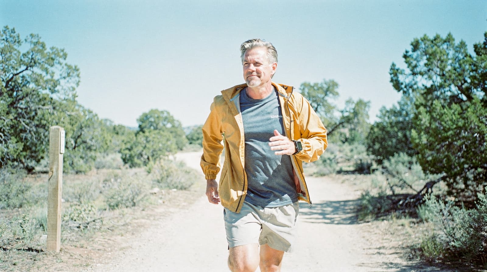 A man in a yellow jacket jogs on a dirt path surrounded by trees and shrubs under a clear blue sky.