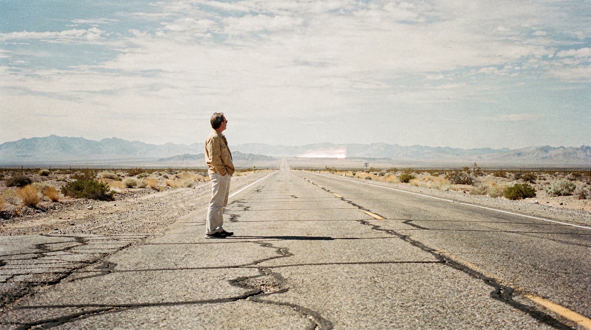 A man stands on a cracked, deserted road in a desert landscape with mountains in the distance.