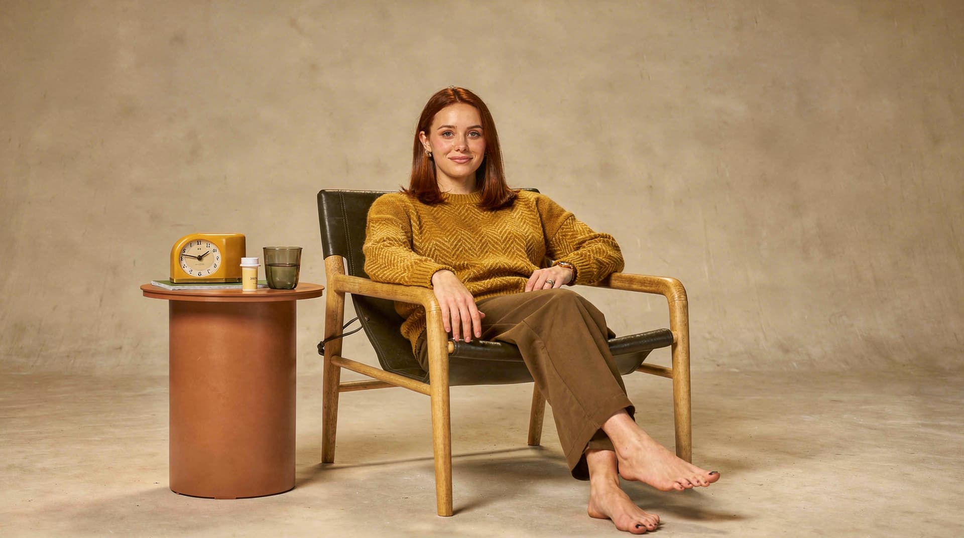 A woman in a yellow sweater sits barefoot in a chair next to a small table with a clock and a cup.