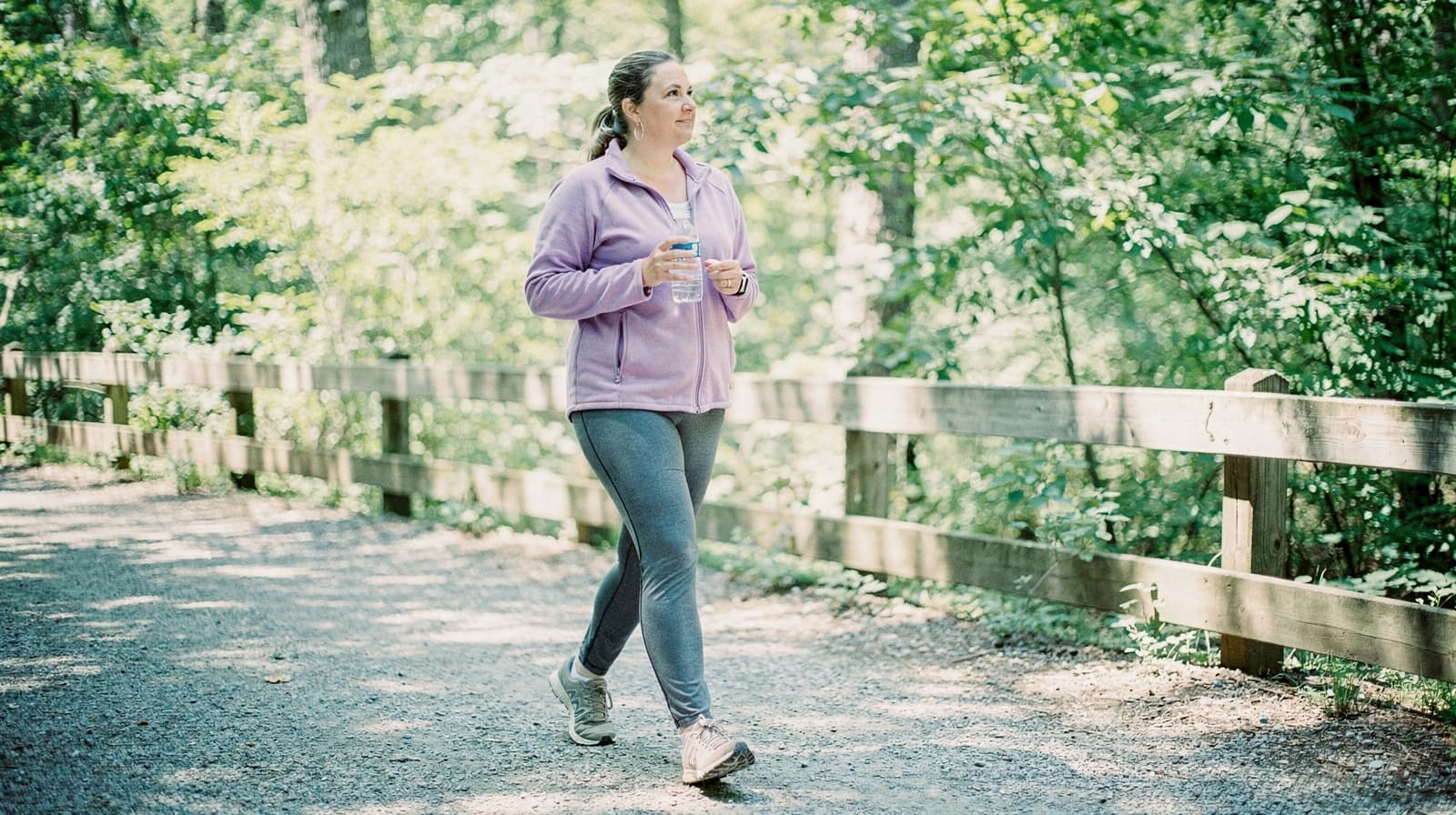 A woman in athletic wear walks on a wooded trail holding a water bottle.