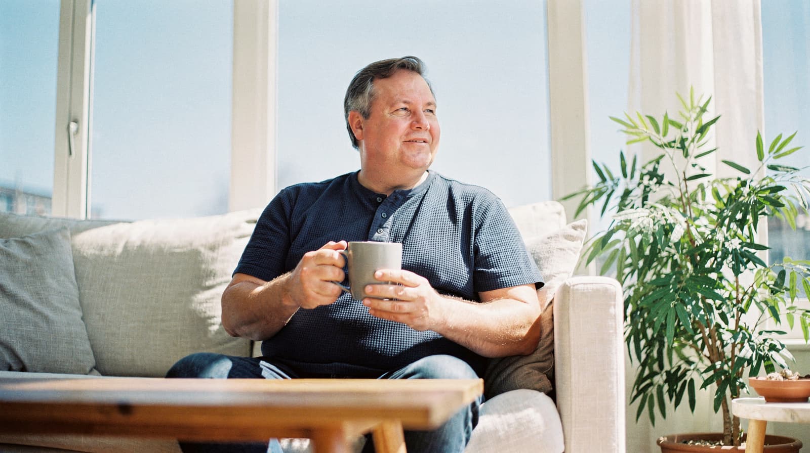 A man sits on a sofa holding a mug, looking out a window with sunlight streaming in, next to a potted plant.