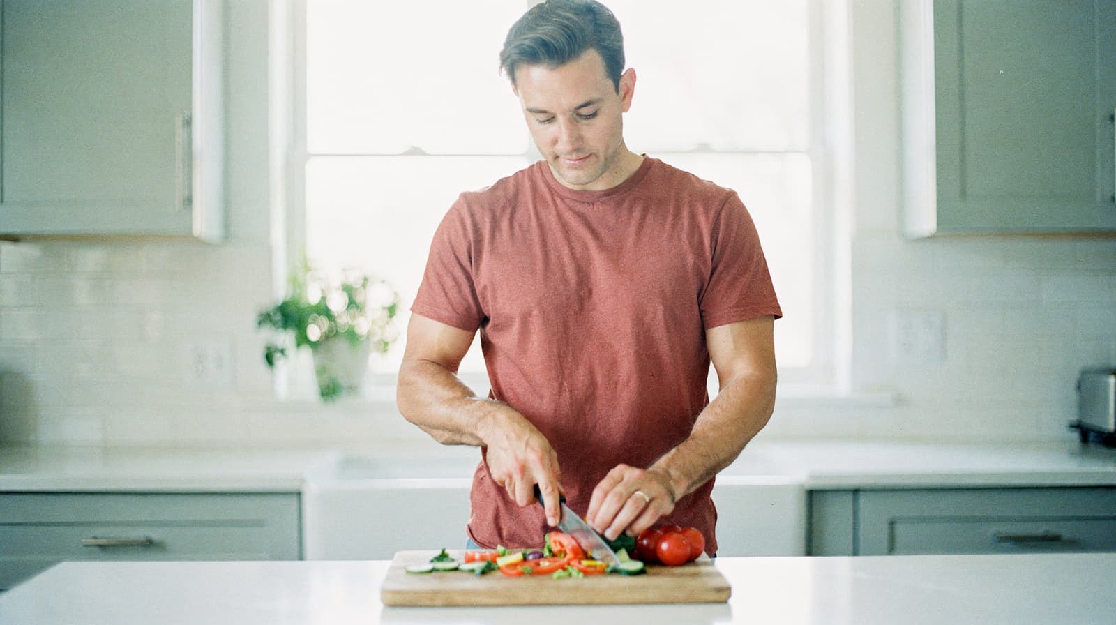A man in a red shirt slices vegetables on a cutting board in a bright kitchen.