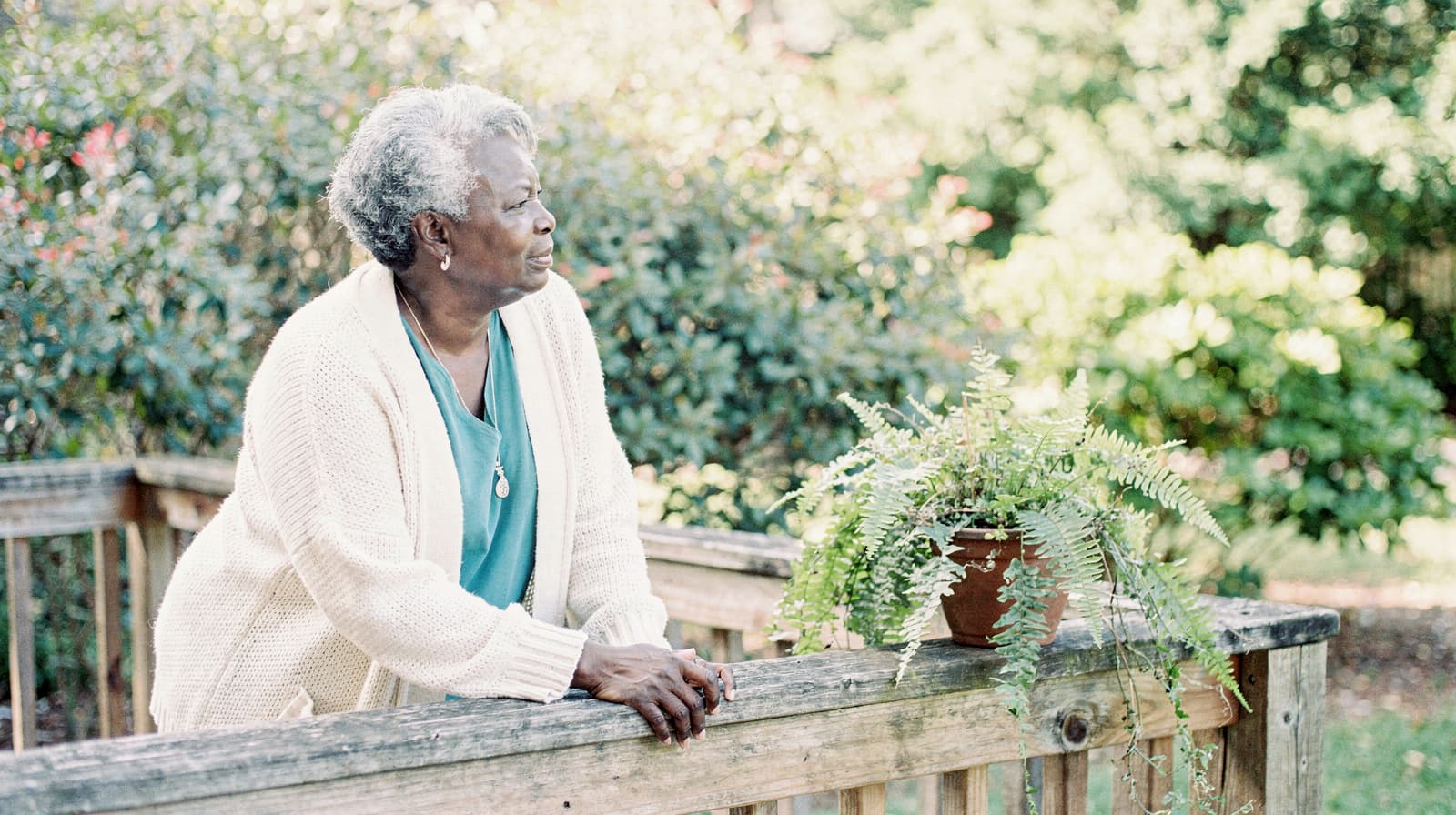 A woman in a white sweater leans on a wooden railing next to a potted fern, with greenery in the background.