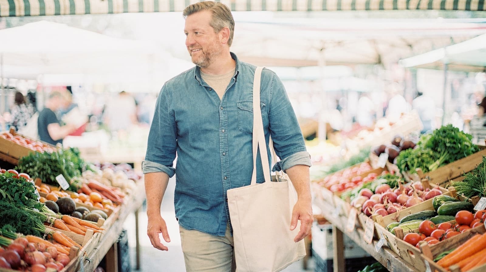 A man carrying a tote bag walks through a farmers market with various fruits and vegetables displayed on wooden stands.