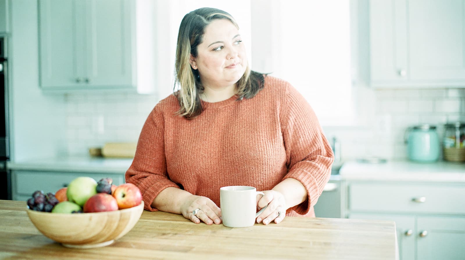 A woman in an orange sweater holds a mug while sitting at a kitchen counter with a bowl of fruit.
