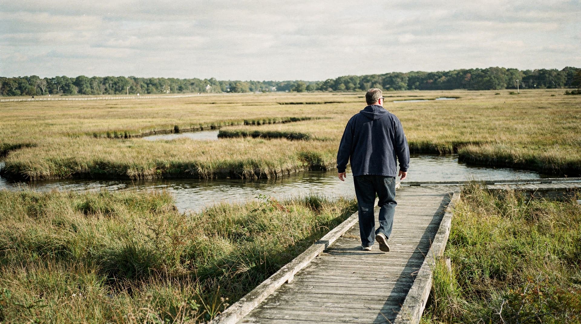 A man walks along a wooden boardwalk through a grassy marshland under a cloudy sky.