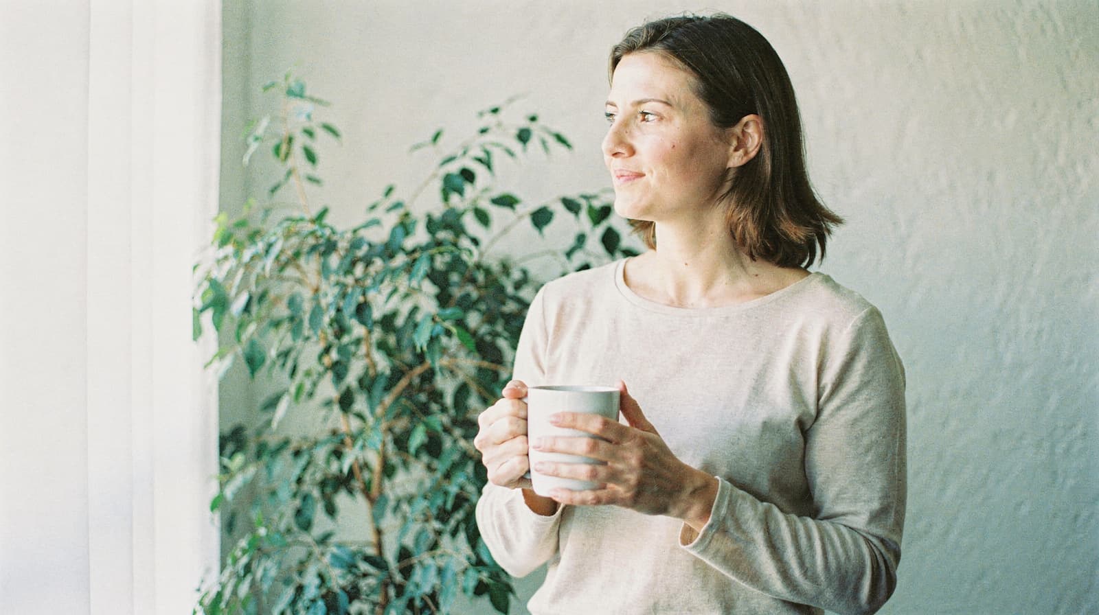A woman holding a mug looks out a window with a potted plant in the background.