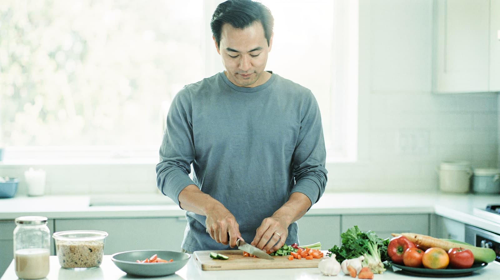 A man chops vegetables on a kitchen counter with various ingredients around him.