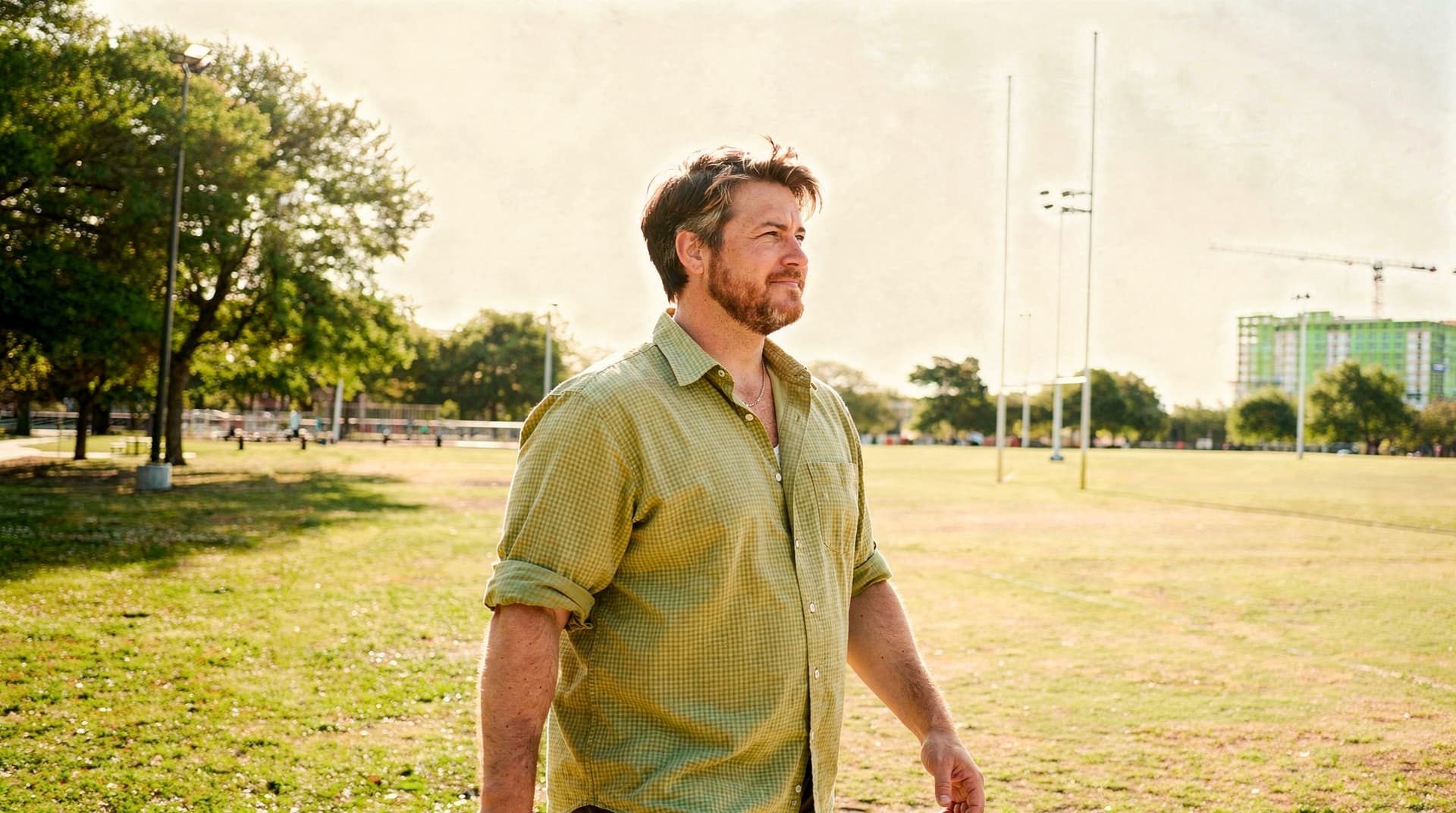 A man in a green shirt walks through a grassy park with trees and buildings in the background.