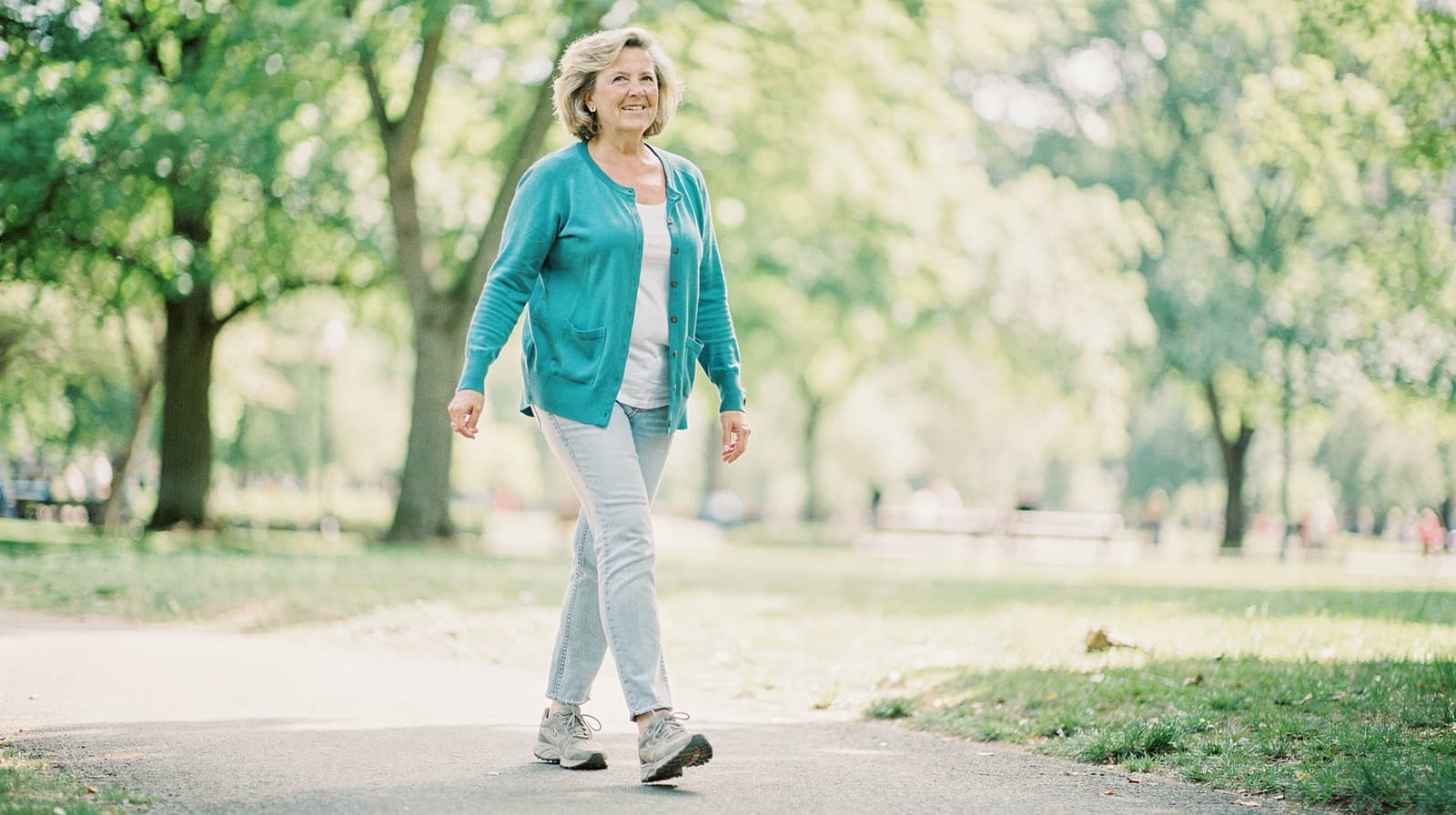 A woman in a blue cardigan walks on a path through a sunlit park with trees in the background.