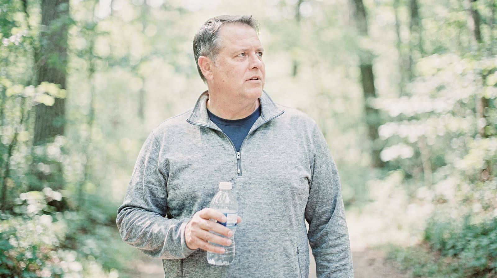 A man holding a water bottle stands on a forest trail, surrounded by trees.