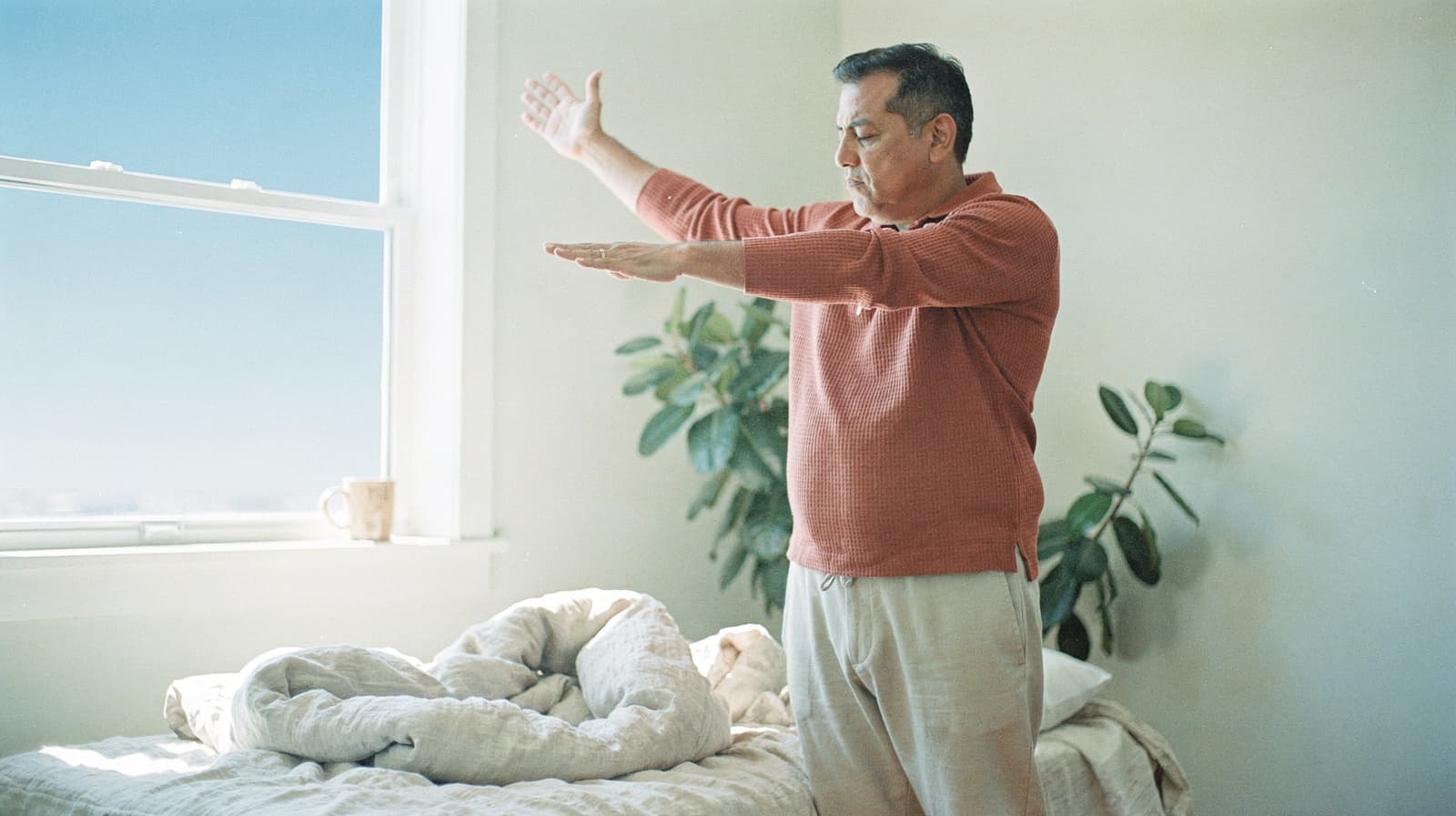A man stands in a bedroom with arms outstretched, next to a bed and a window with a plant nearby.