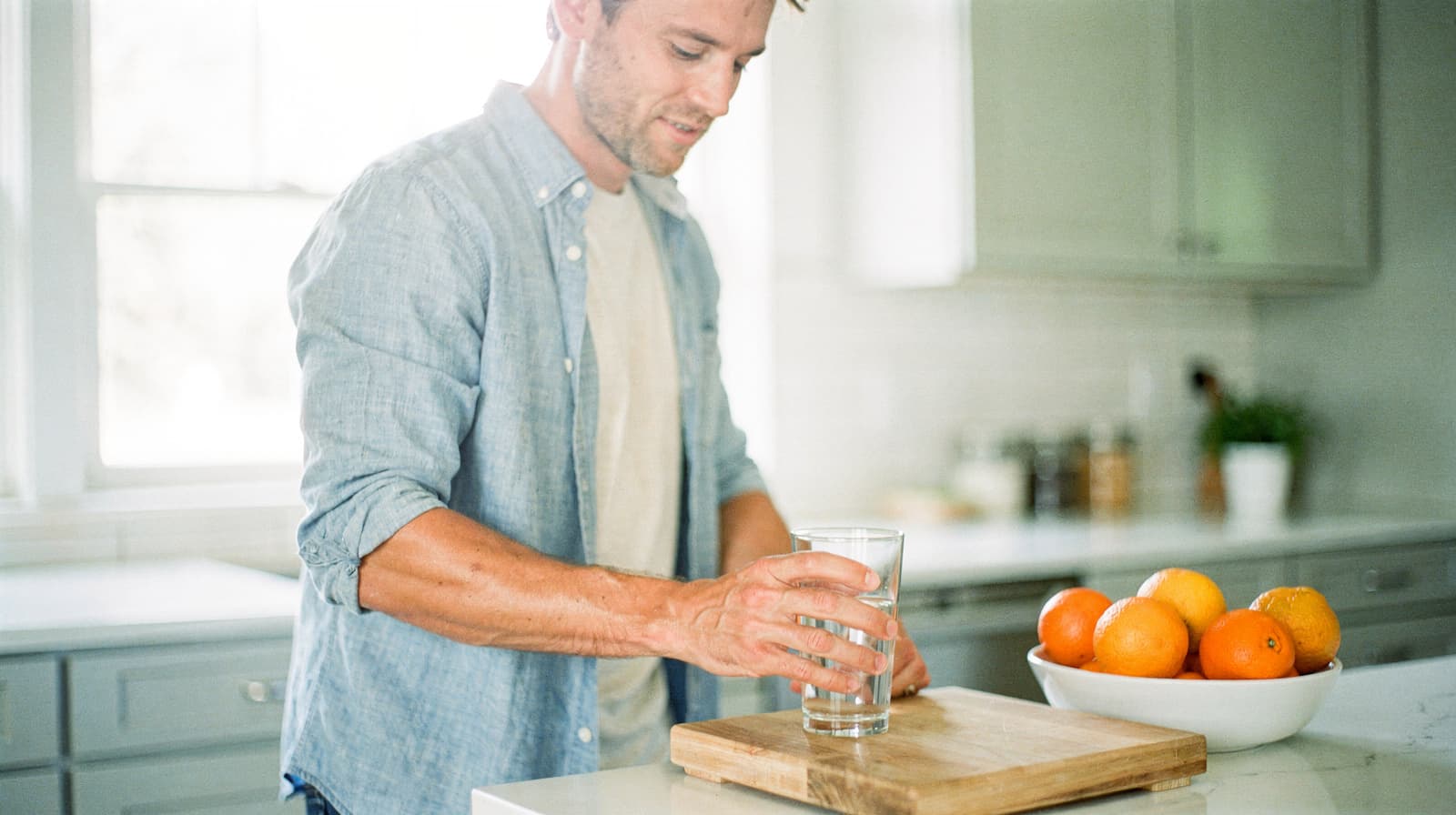 A man in a kitchen holds a glass of water near a bowl of oranges on a countertop.