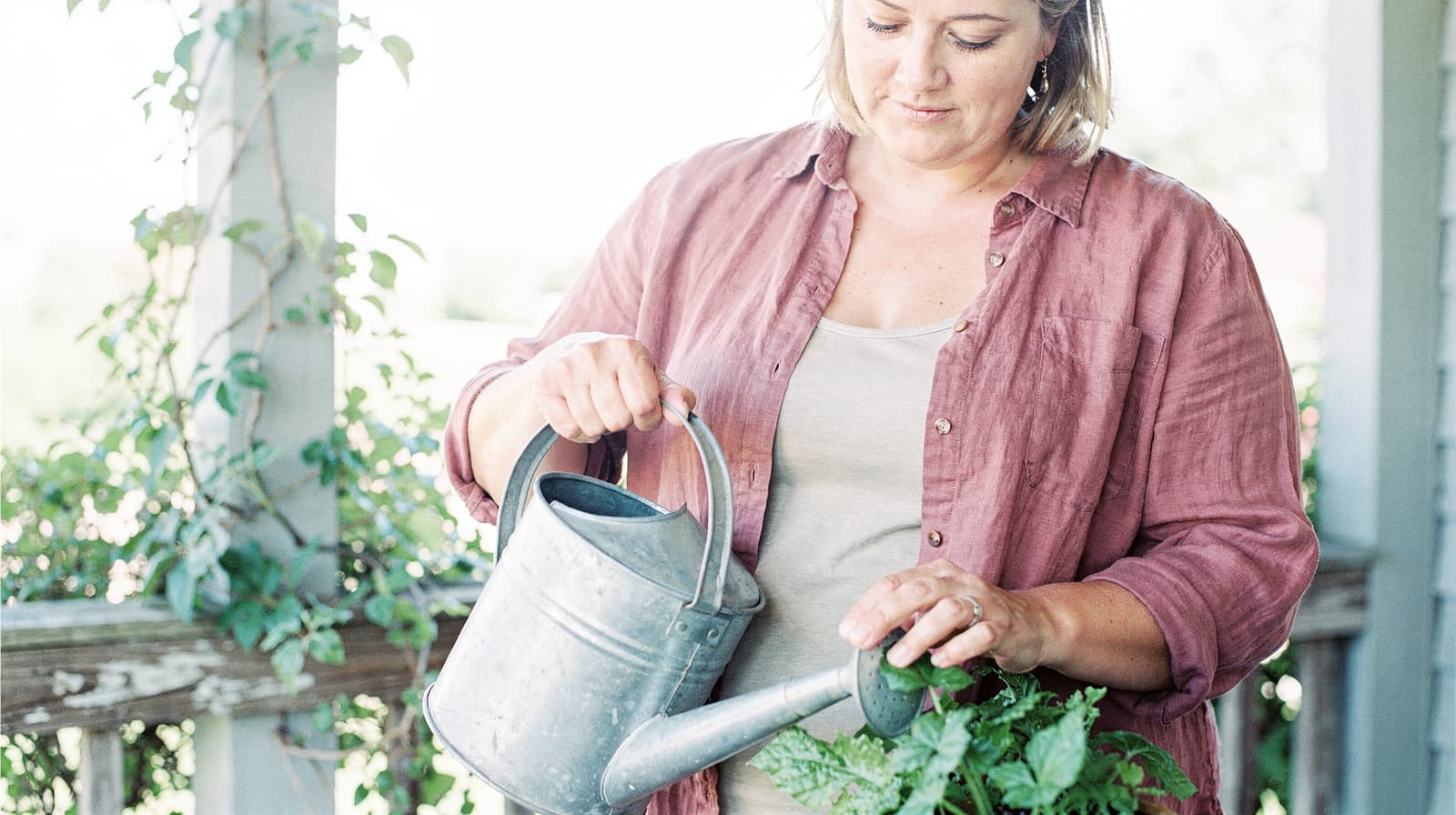 A woman waters plants on a porch with a metal watering can.
