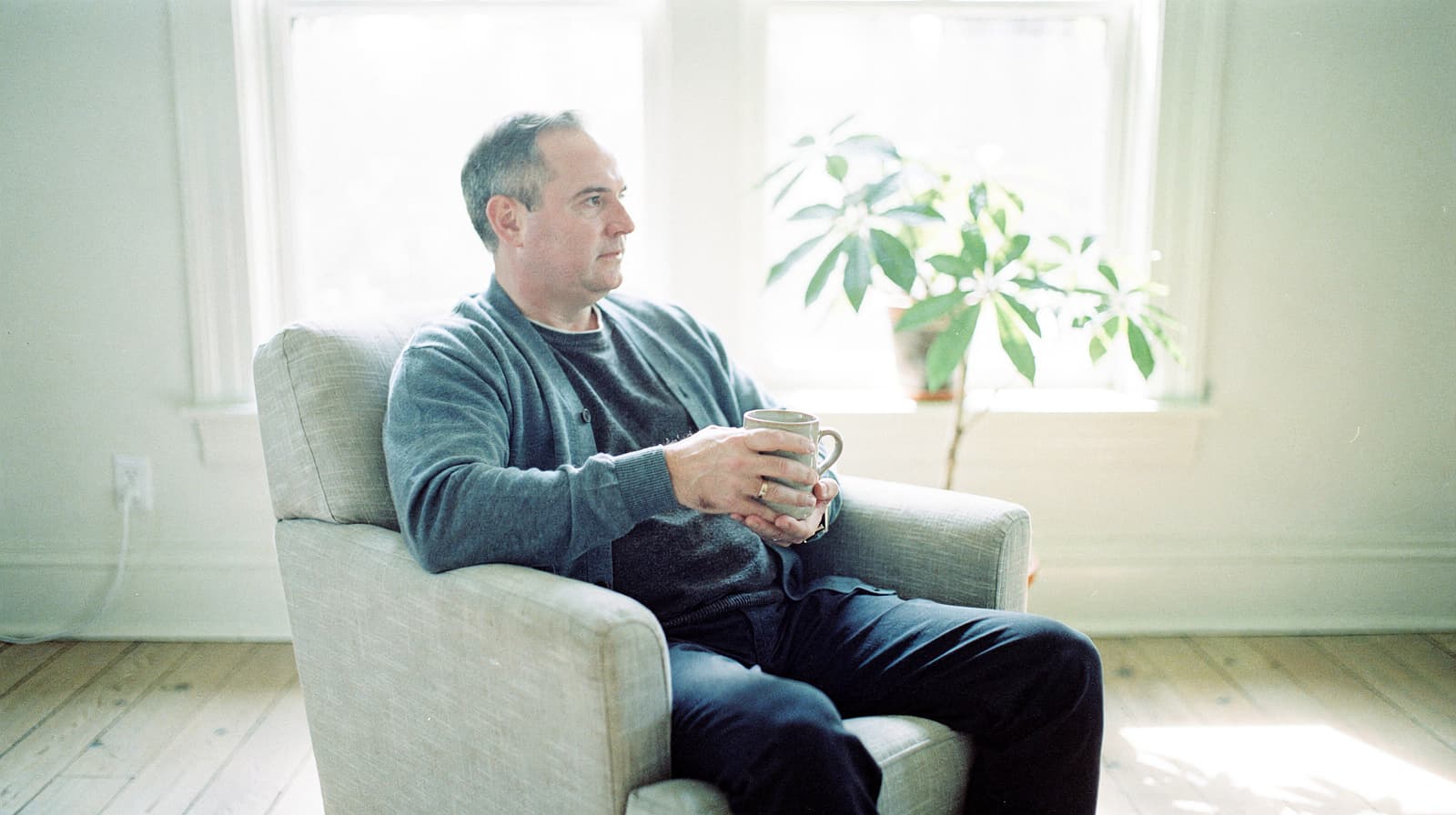 A man sits in an armchair holding a mug, with a plant and two windows in the background.