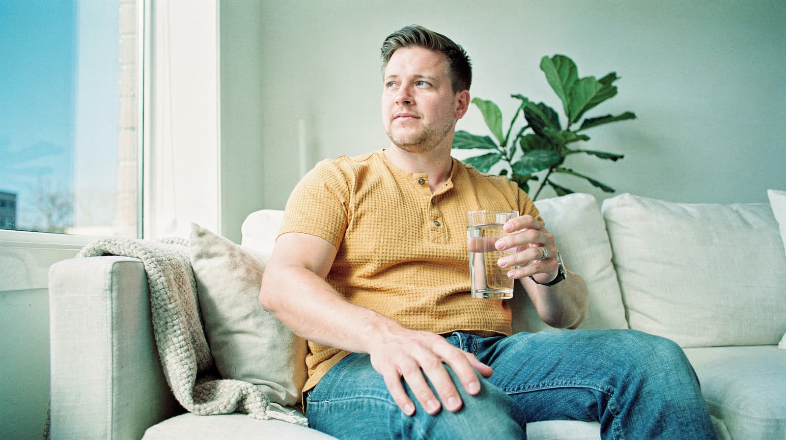 A man in a yellow shirt sits on a sofa holding a glass of water, with a plant in the background.