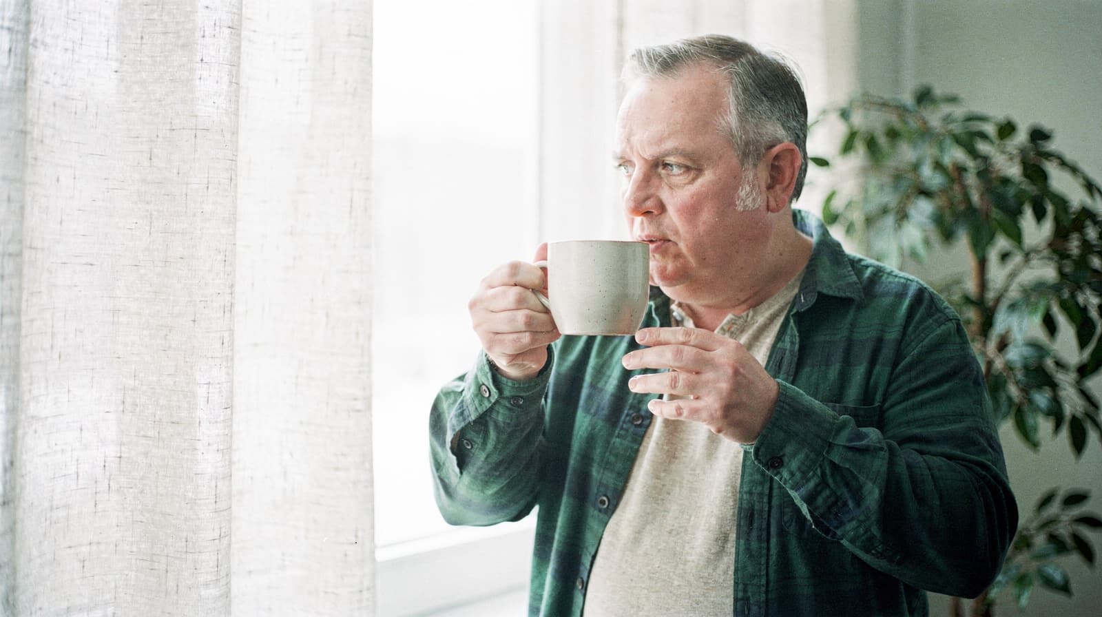 A man in a plaid shirt drinks from a mug while standing by a window with sheer curtains.