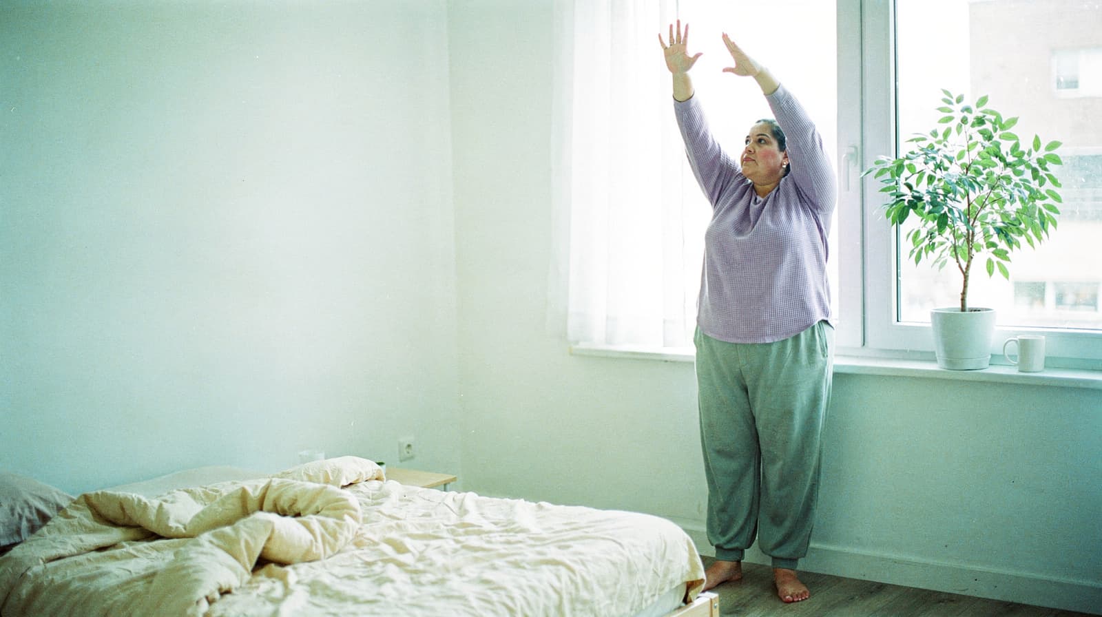 A person in a purple top stretches with arms raised near a window in a bedroom with a bed and a potted plant.