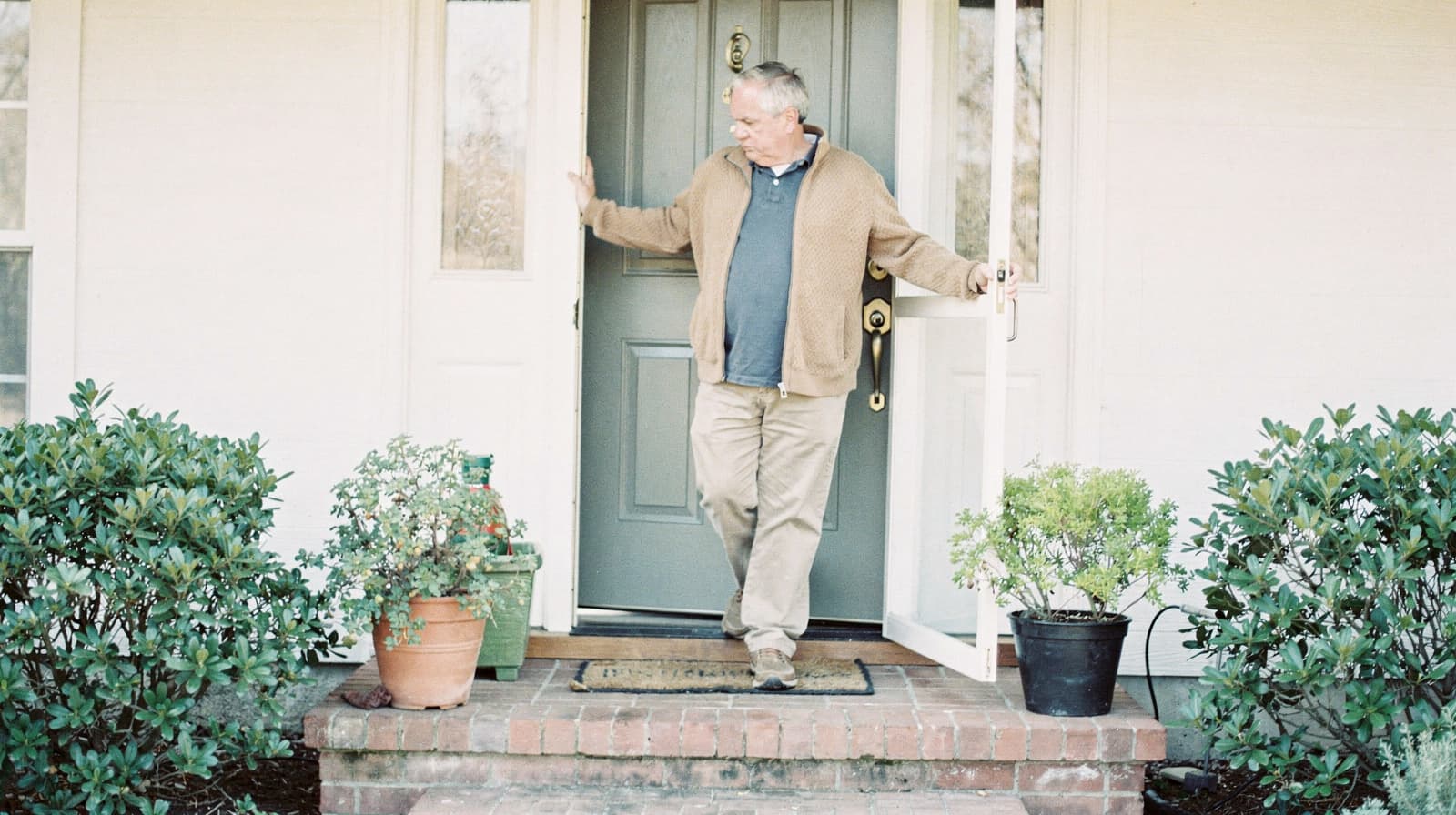 A man stands in the doorway of a house, holding the door open, surrounded by potted plants on a brick step.