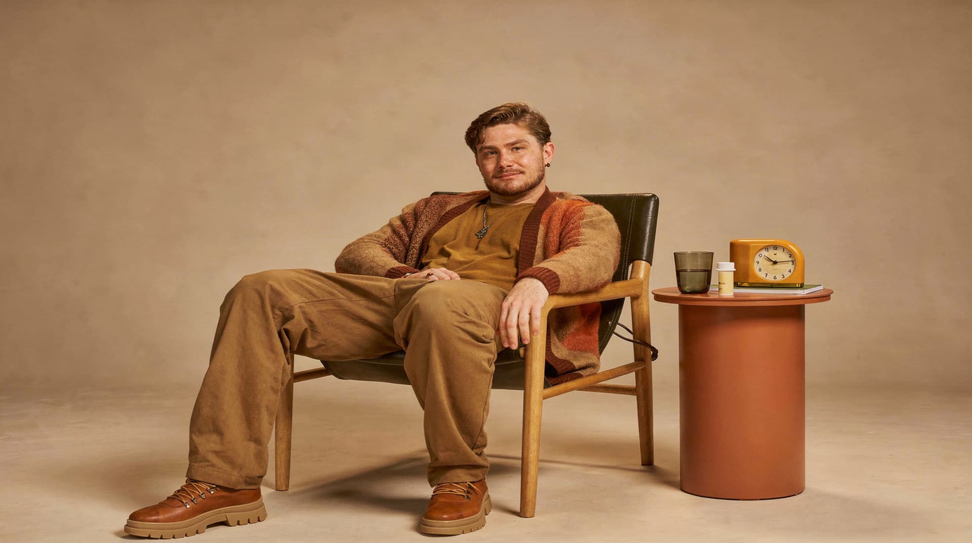 A man in casual clothing sits in a chair next to a small table with a clock and a glass in a neutral-toned room.