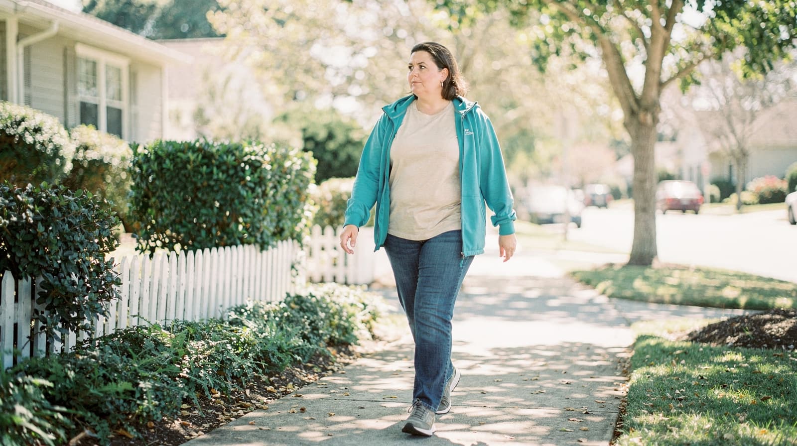 A woman walks on a sunny sidewalk in a suburban neighborhood with trees and houses in the background.