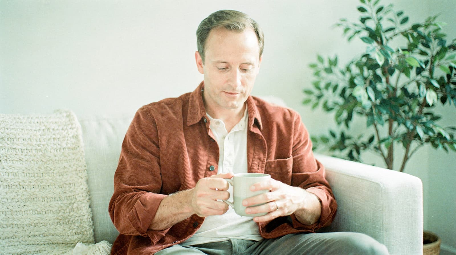 A man sits on a couch holding a mug, with a plant in the background.