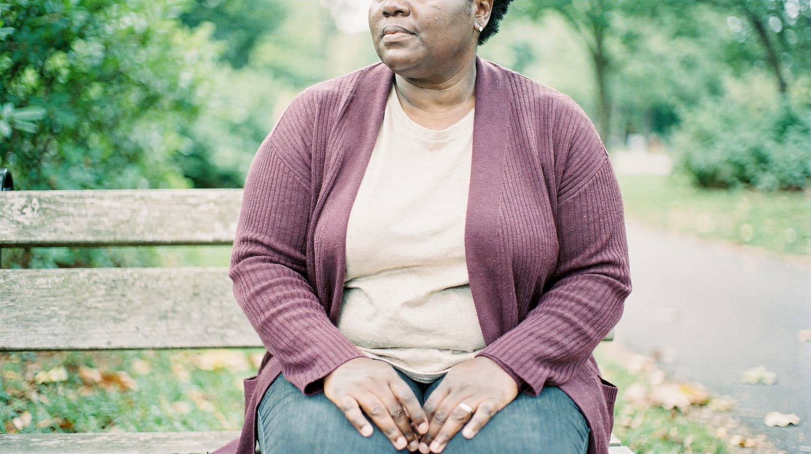 A person sits on a park bench wearing a maroon cardigan and beige shirt, surrounded by green trees.