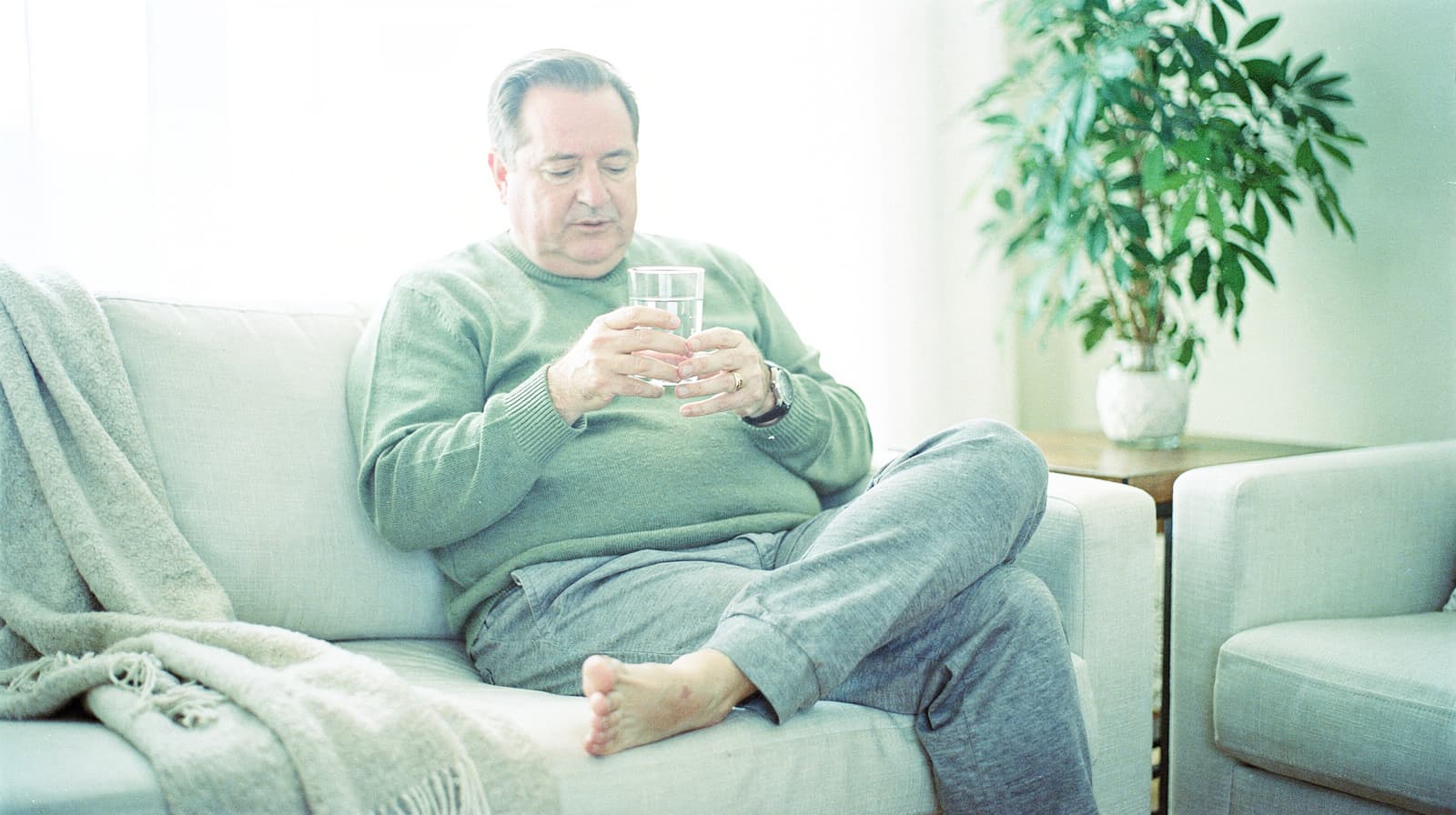 A man in a green sweater sits on a couch holding a glass of water, with a plant in the background.