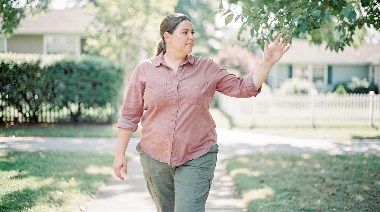 A woman walks on a sidewalk, touching leaves from a tree, with houses and a white fence in the background.