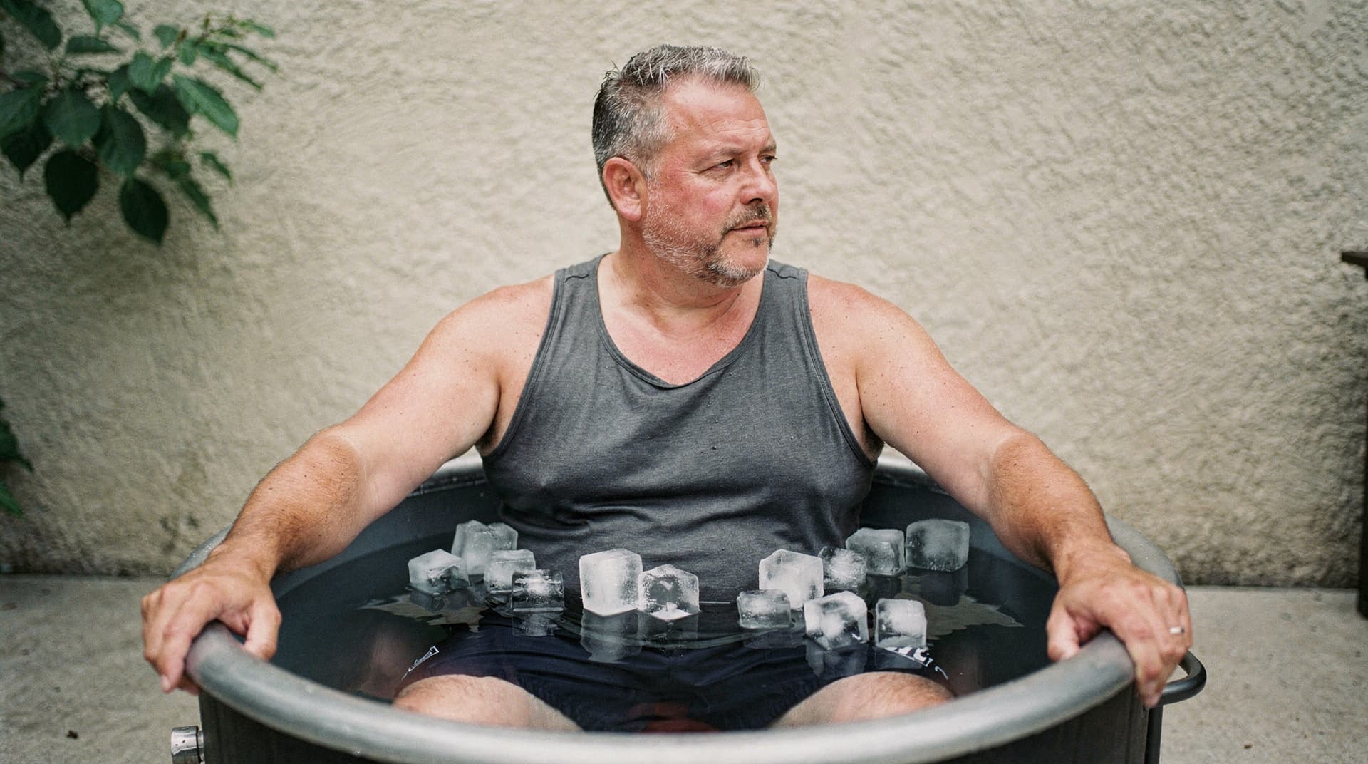 A man sits in an ice bath outdoors, looking to the side with a neutral expression.