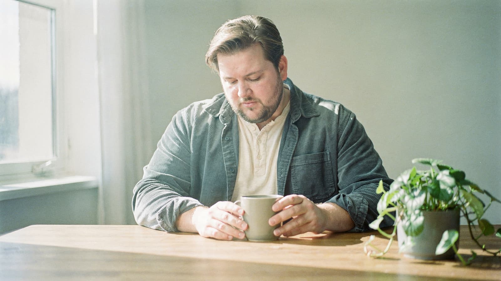 A man sits at a wooden table holding a mug, with a potted plant nearby and sunlight streaming through a window.