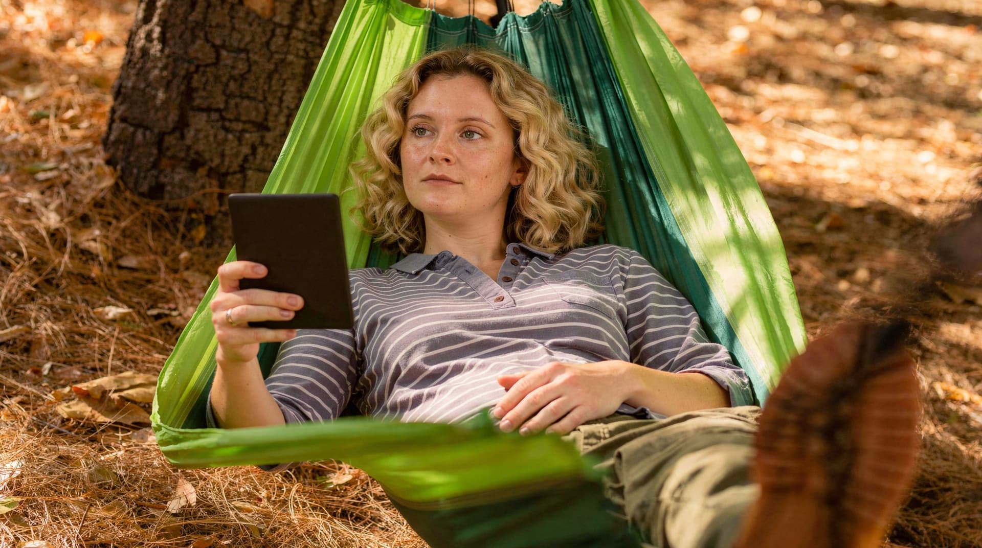 A woman relaxes in a green hammock outdoors, reading a tablet under a tree.