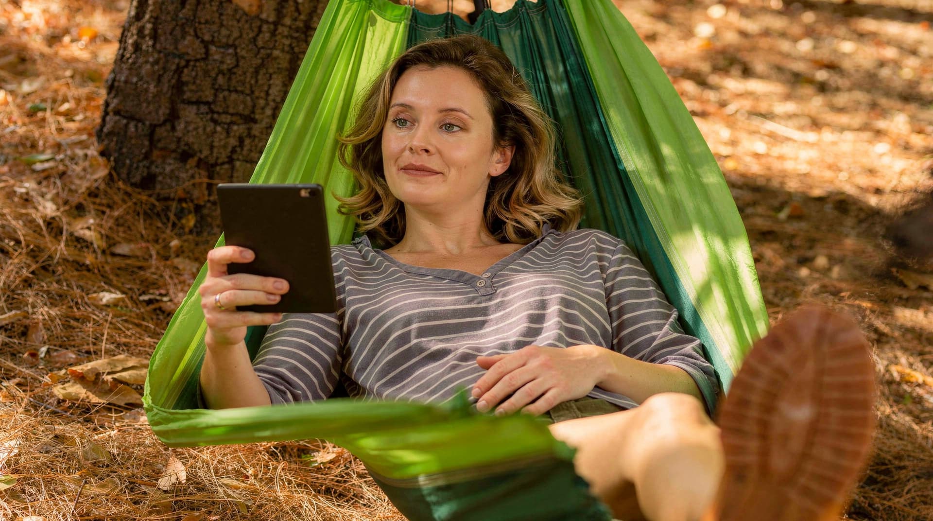 A woman relaxes in a green hammock outdoors, reading a tablet under a tree.