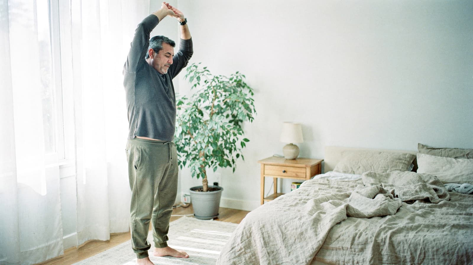 A man stretches with his arms overhead in a bright bedroom next to a bed and a potted plant.