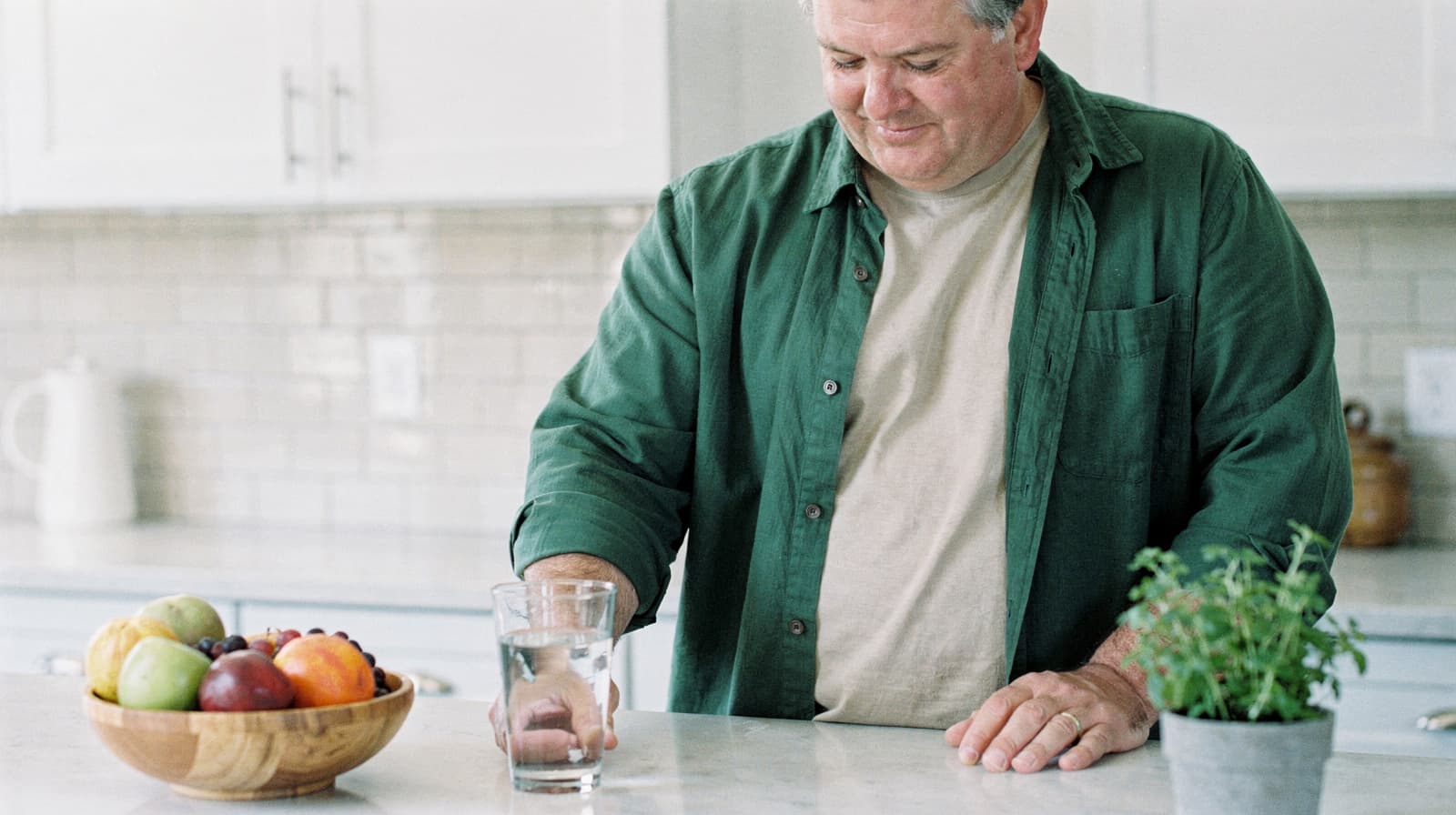 A man in a green shirt stands at a kitchen counter with a glass of water and a bowl of fruit.