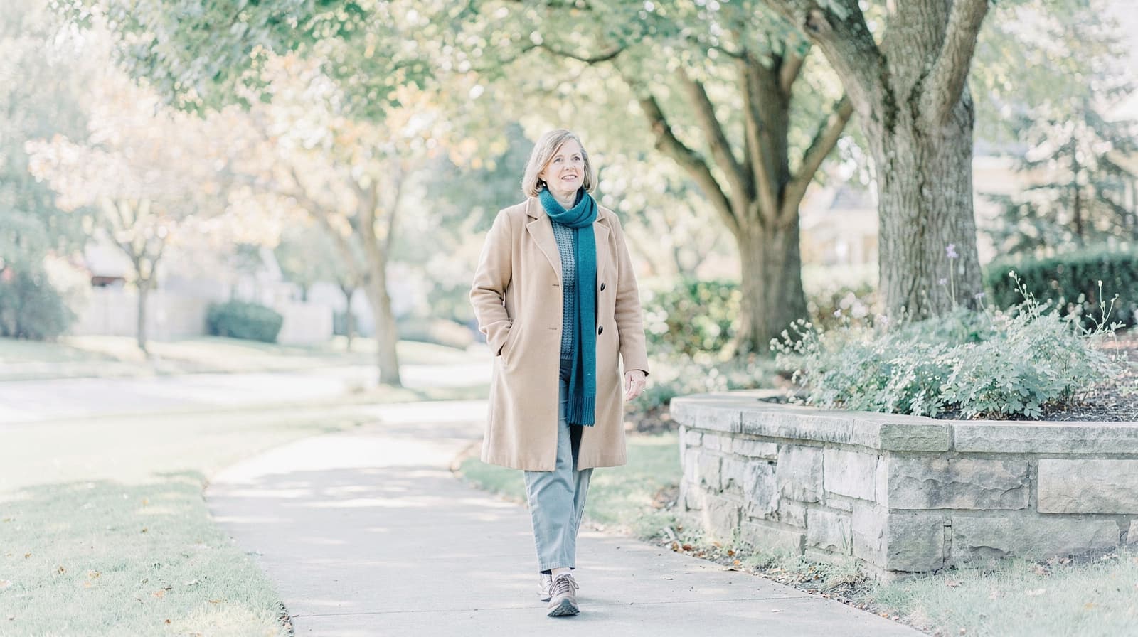 A woman in a coat walks along a tree-lined sidewalk on a sunny day.