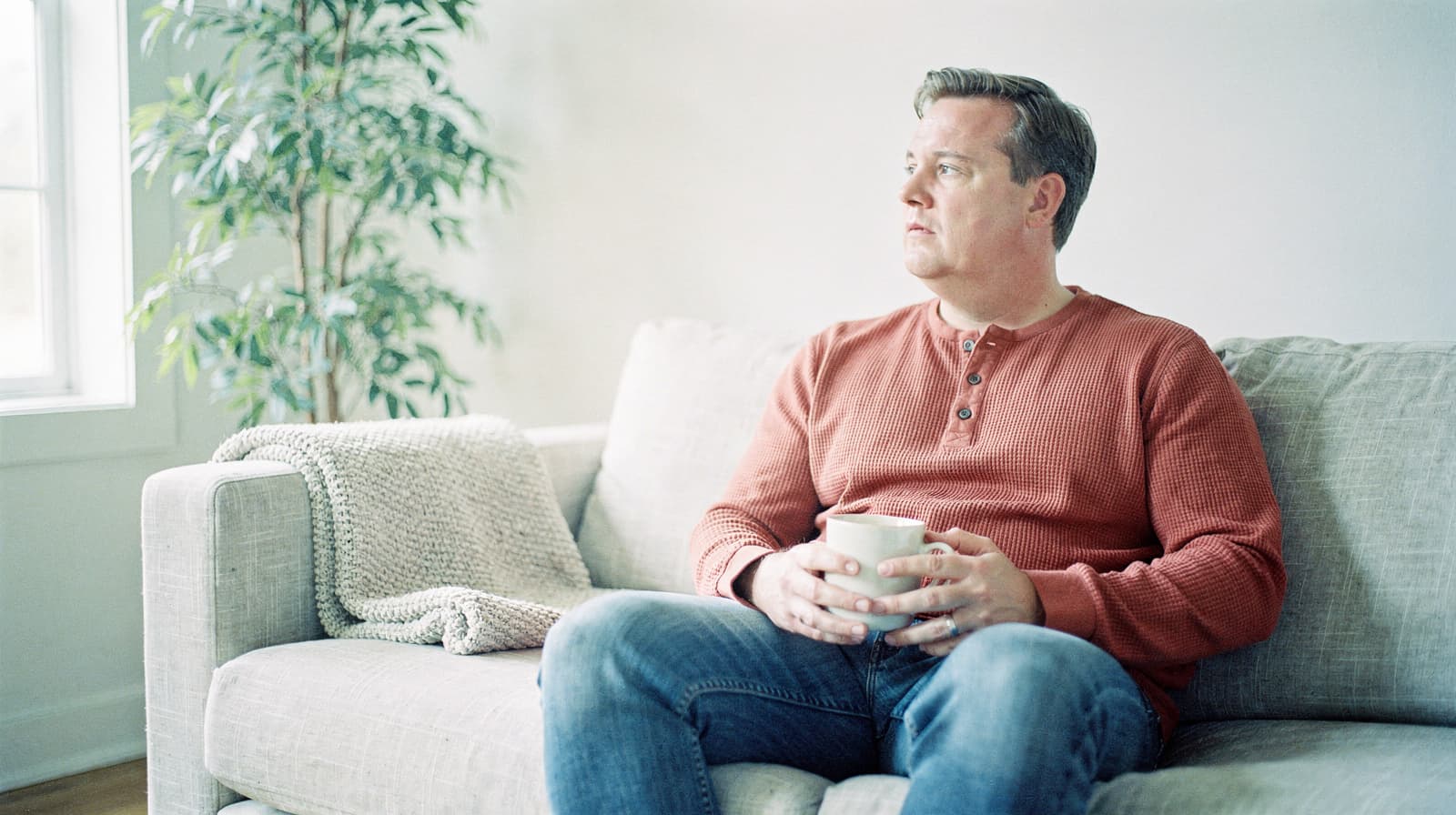 A man sits on a sofa holding a mug, looking out a window with a plant nearby.