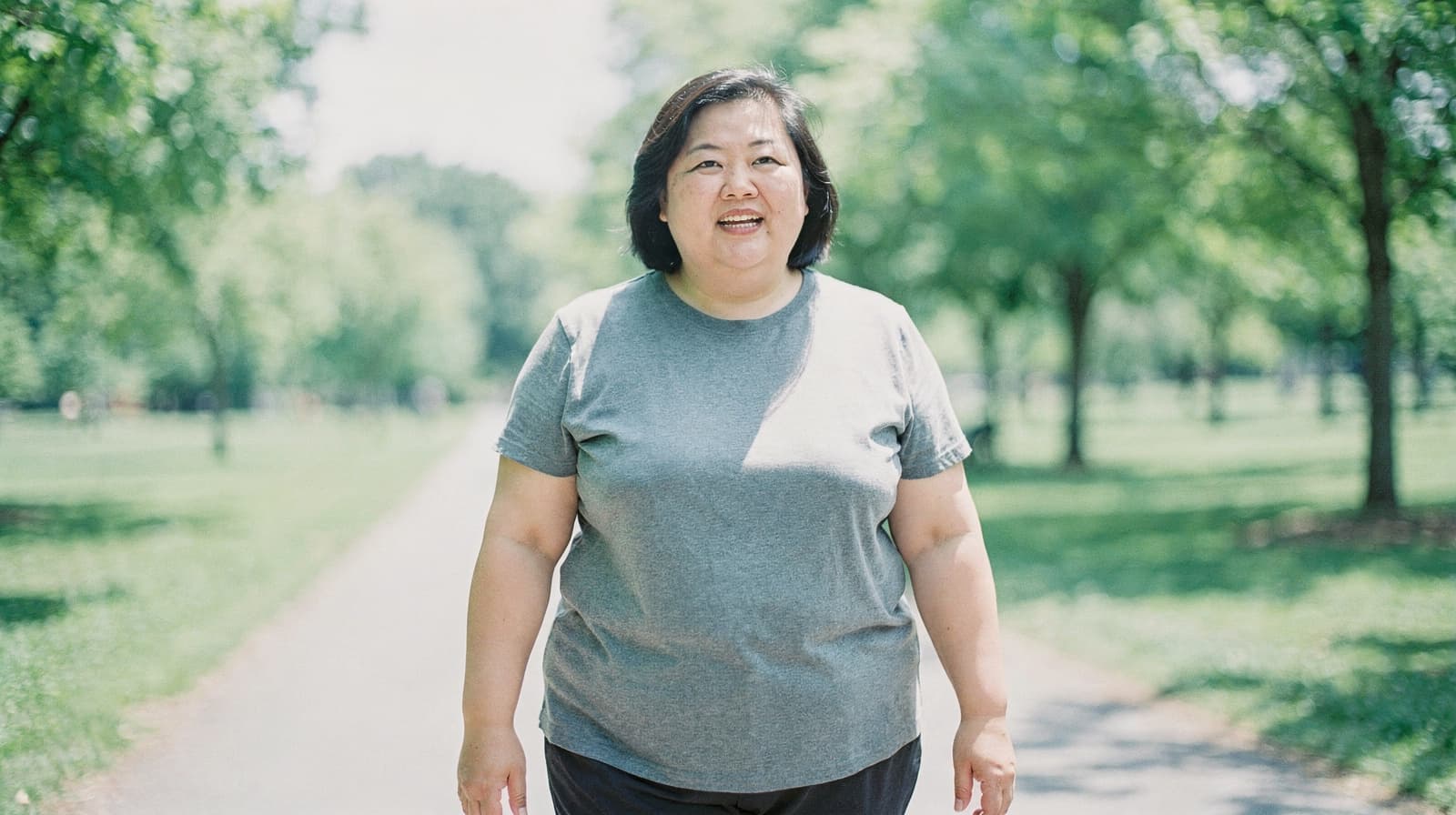 A woman walks along a tree-lined path in a sunny park.
