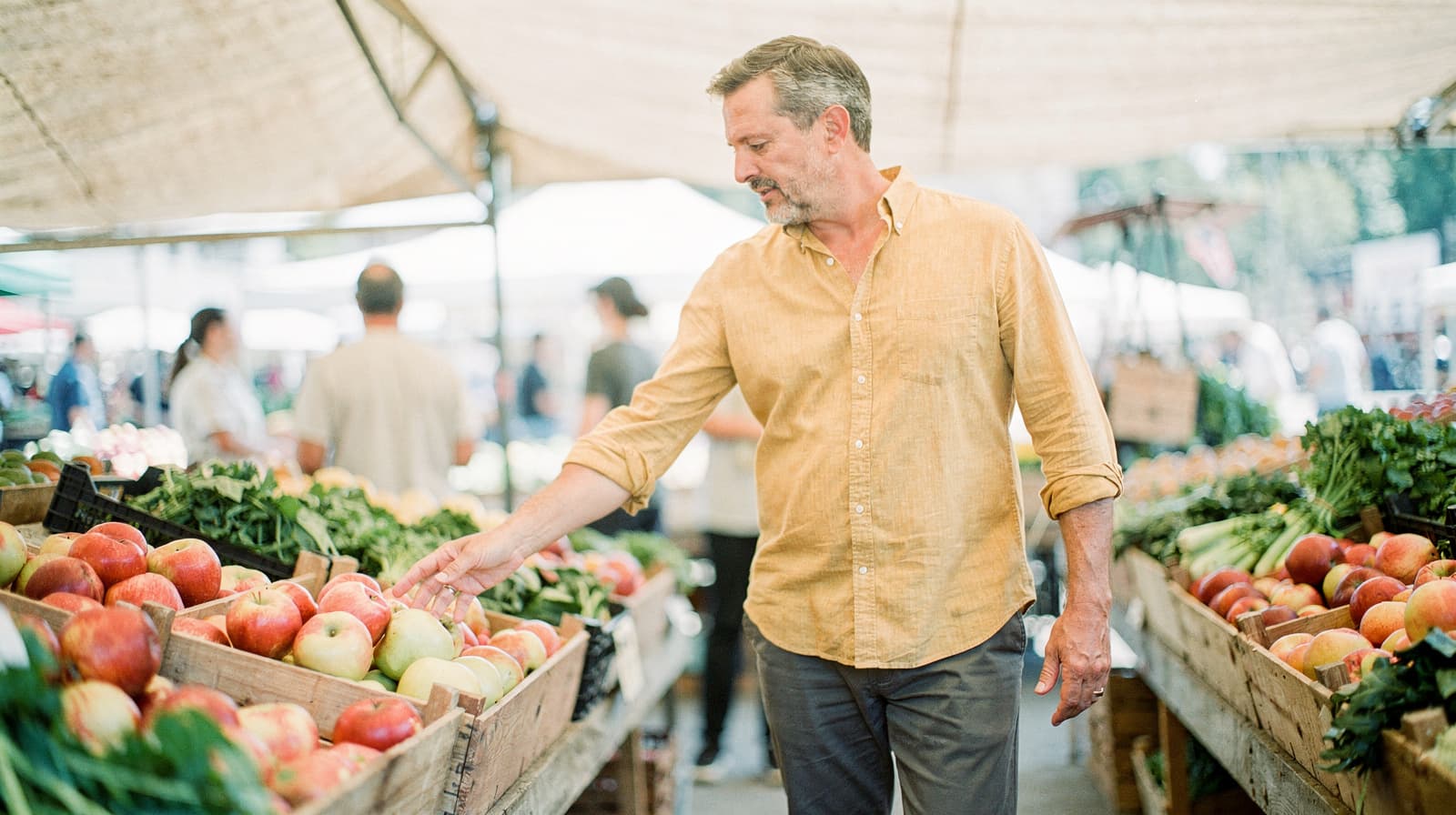 A man in a yellow shirt selects apples at an outdoor market stall.