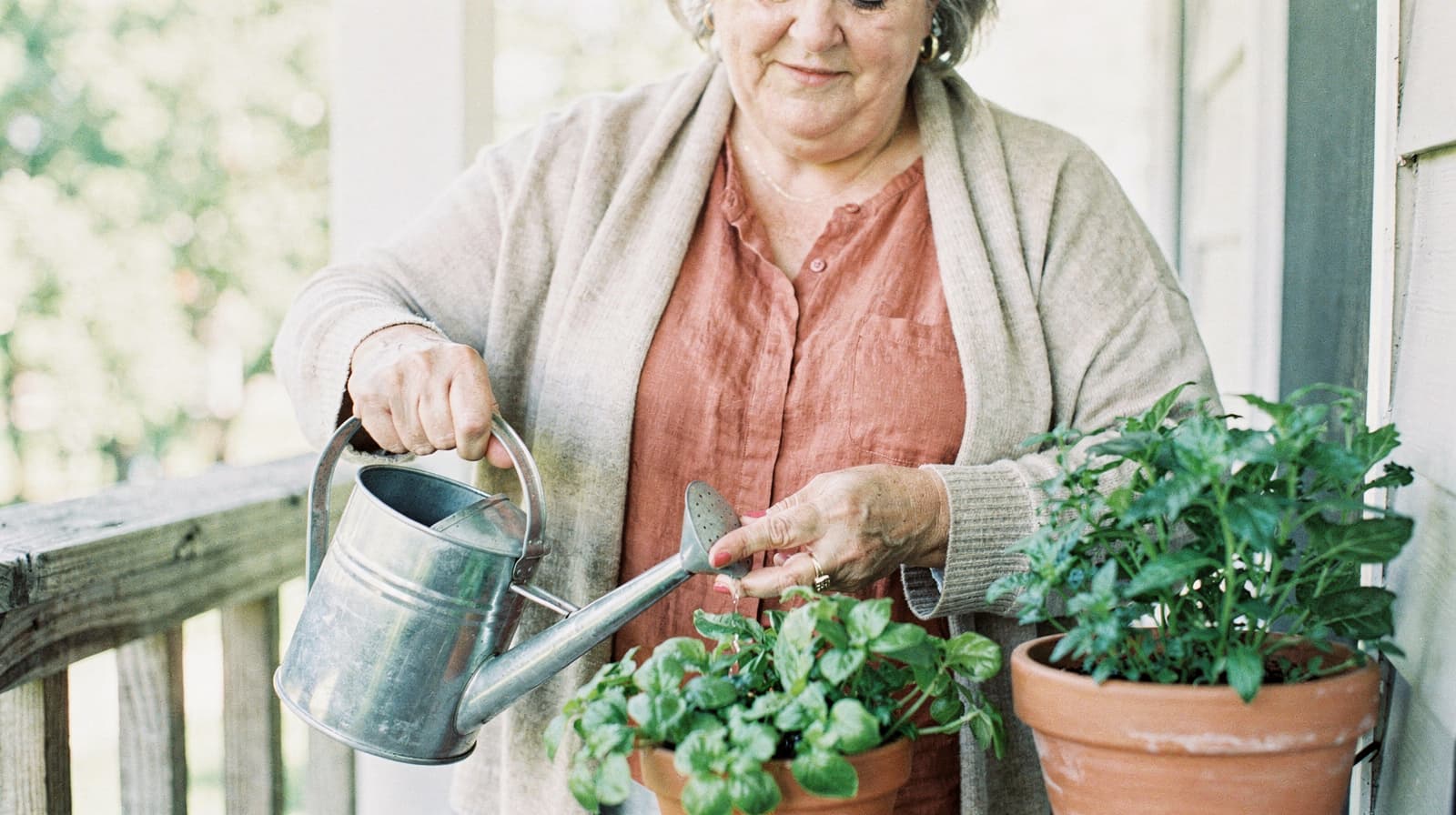 A woman waters potted plants on a porch using a metal watering can.