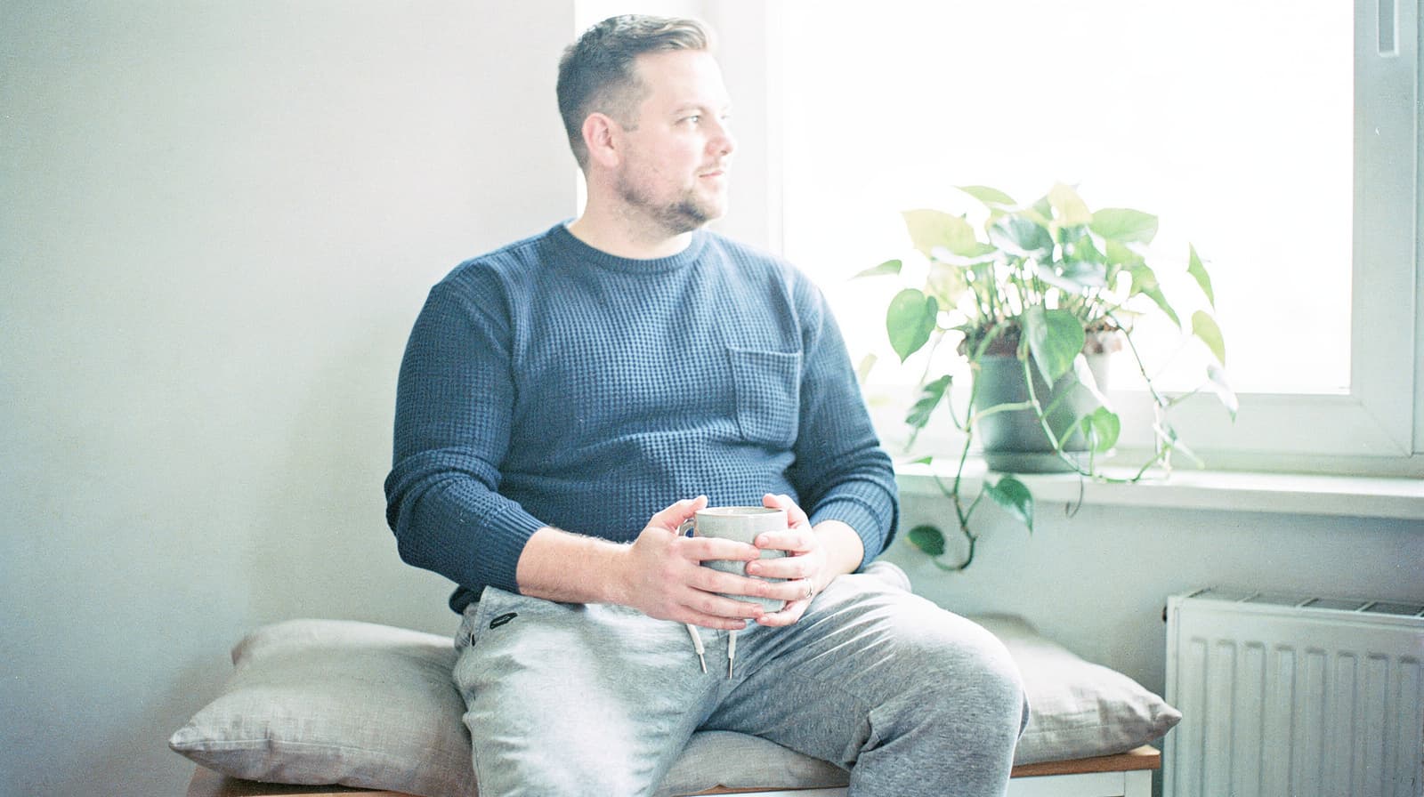 A man sits on a bench holding a mug, looking out a window with a potted plant nearby.