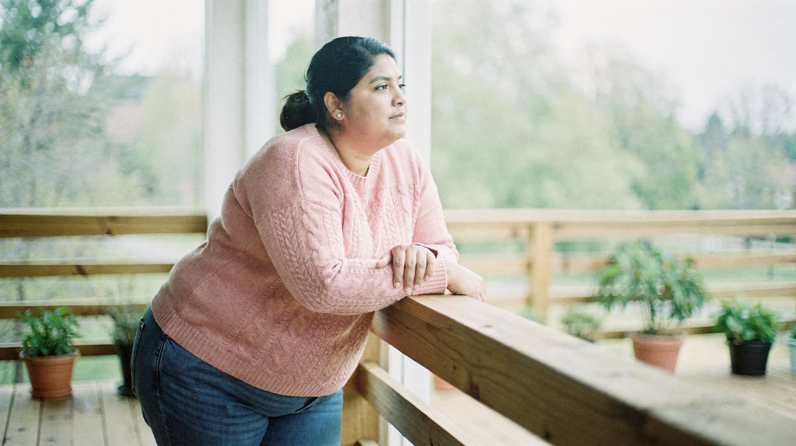 A woman in a pink sweater leans on a wooden railing, looking out at a garden with potted plants.