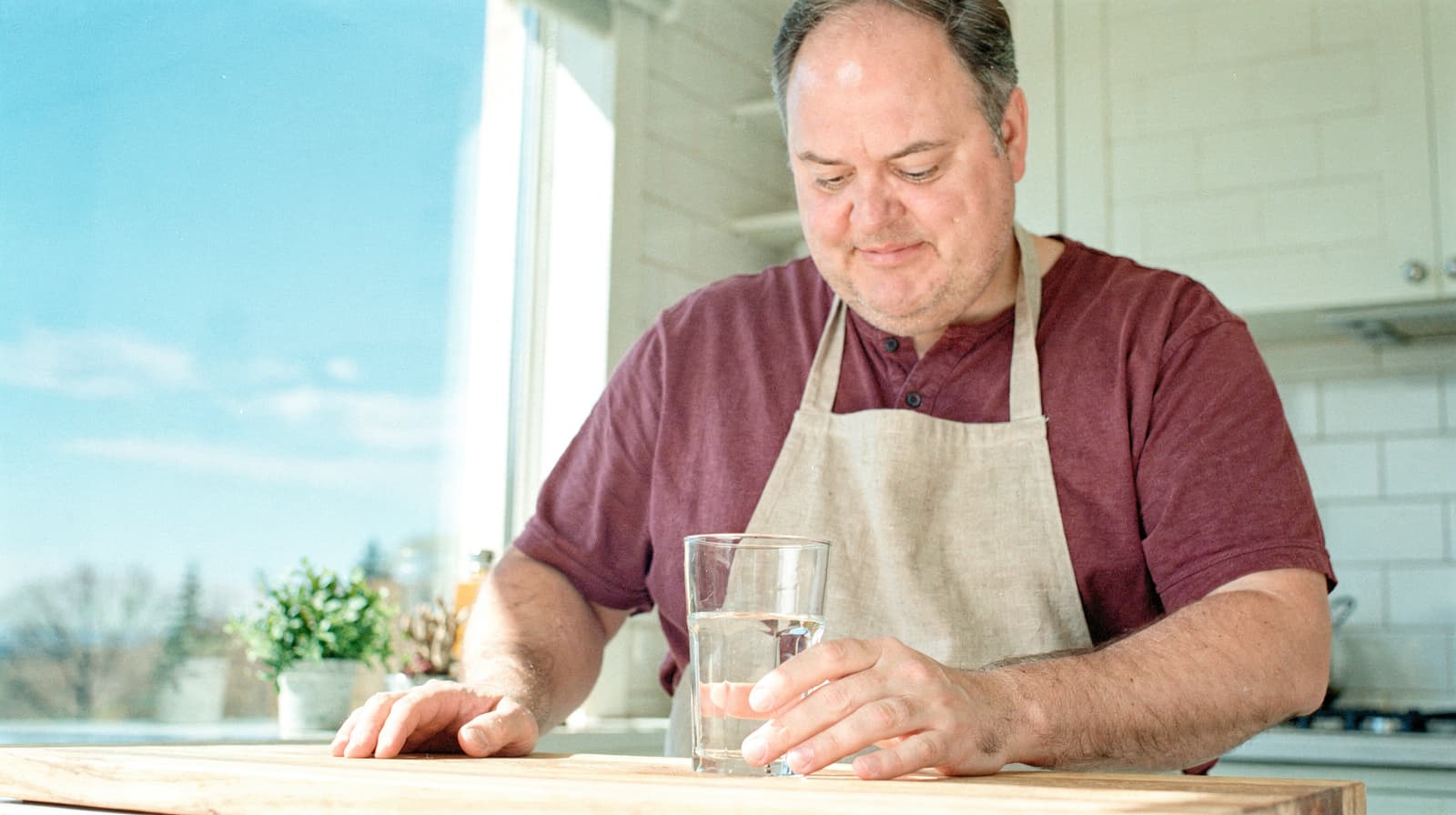 A man in a red shirt and apron holds a glass of water at a kitchen counter with a window view.