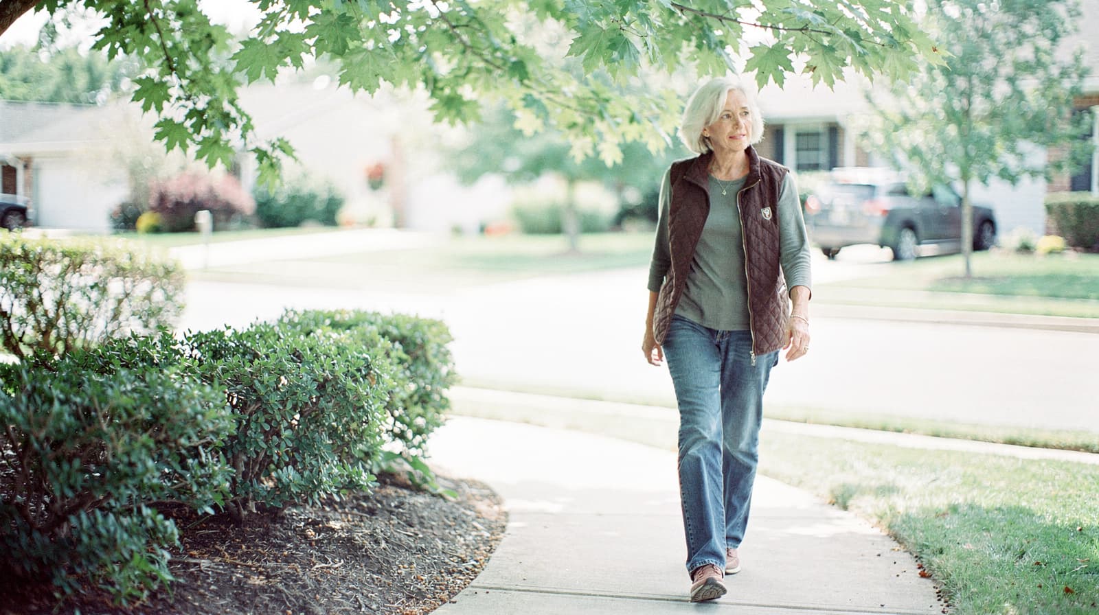 A woman walks on a sidewalk under a tree in a suburban neighborhood.