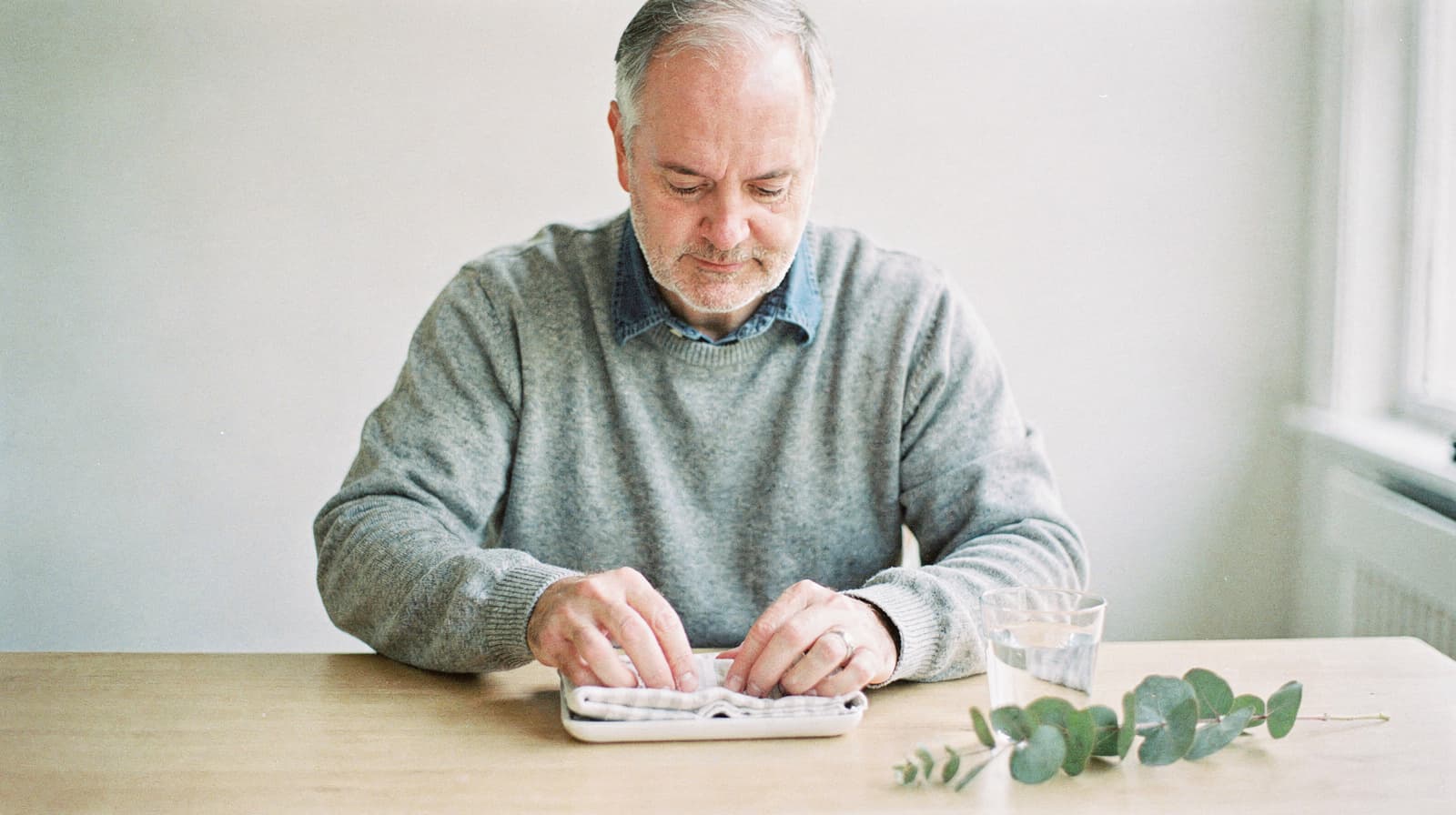 A man sits at a table organizing pills into a pill organizer with a glass of water and eucalyptus leaves nearby.