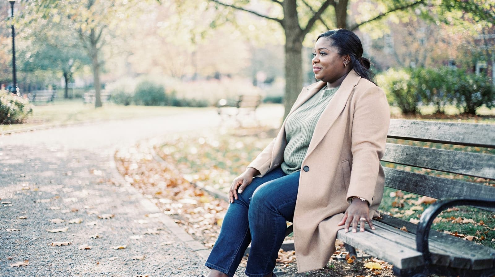 A woman in a coat sits on a park bench surrounded by autumn leaves.