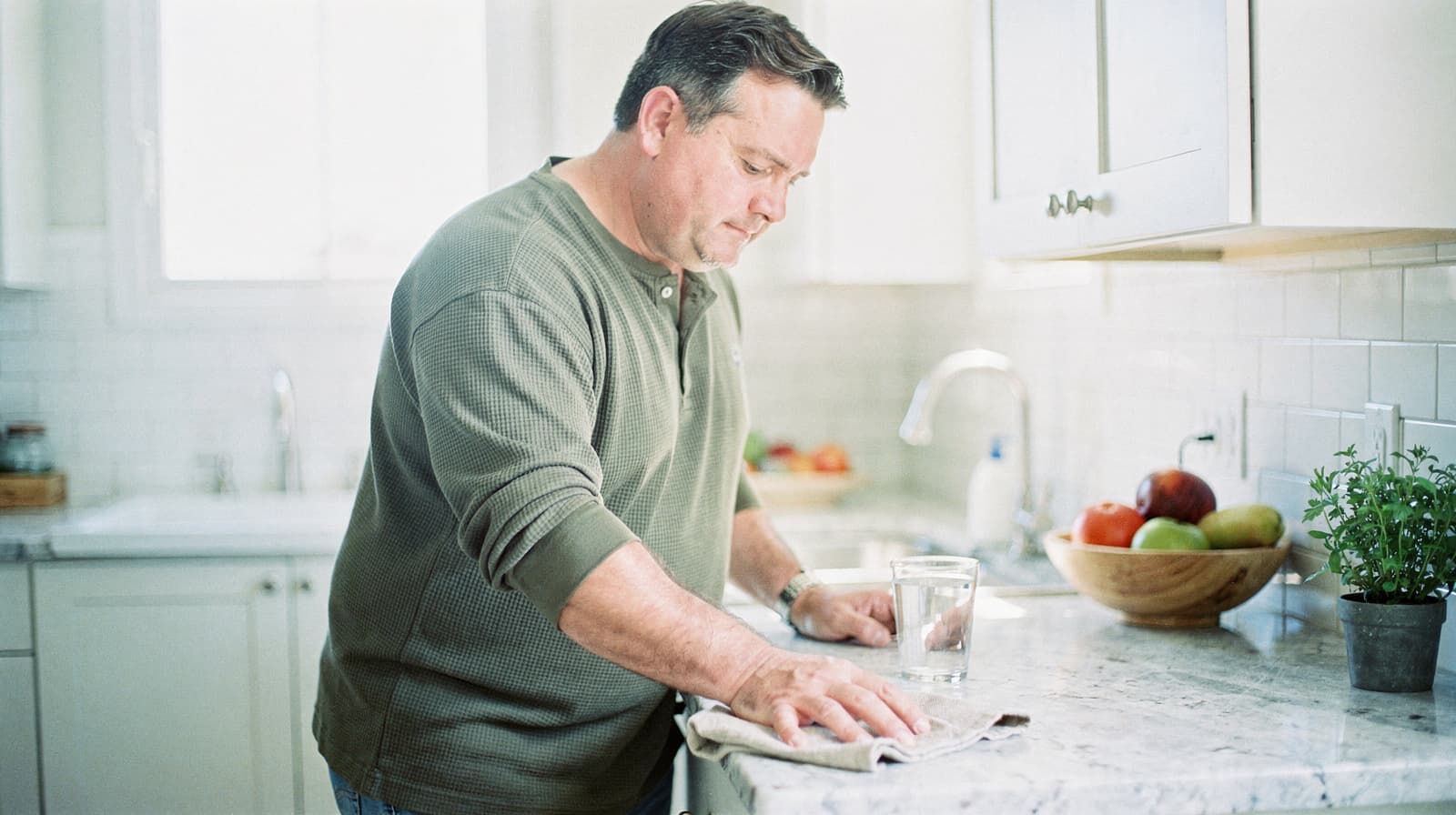 A man wipes a kitchen counter with a cloth next to a glass of water and a bowl of fruit.