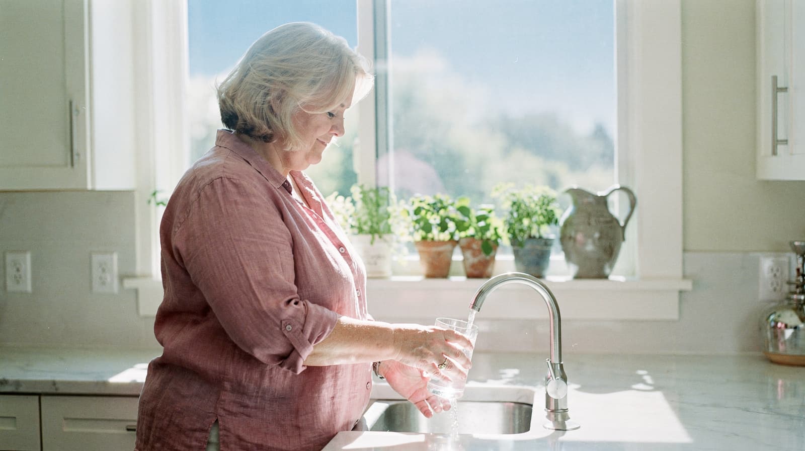 A woman fills a glass with water at a kitchen sink, with potted plants on the windowsill in the background.