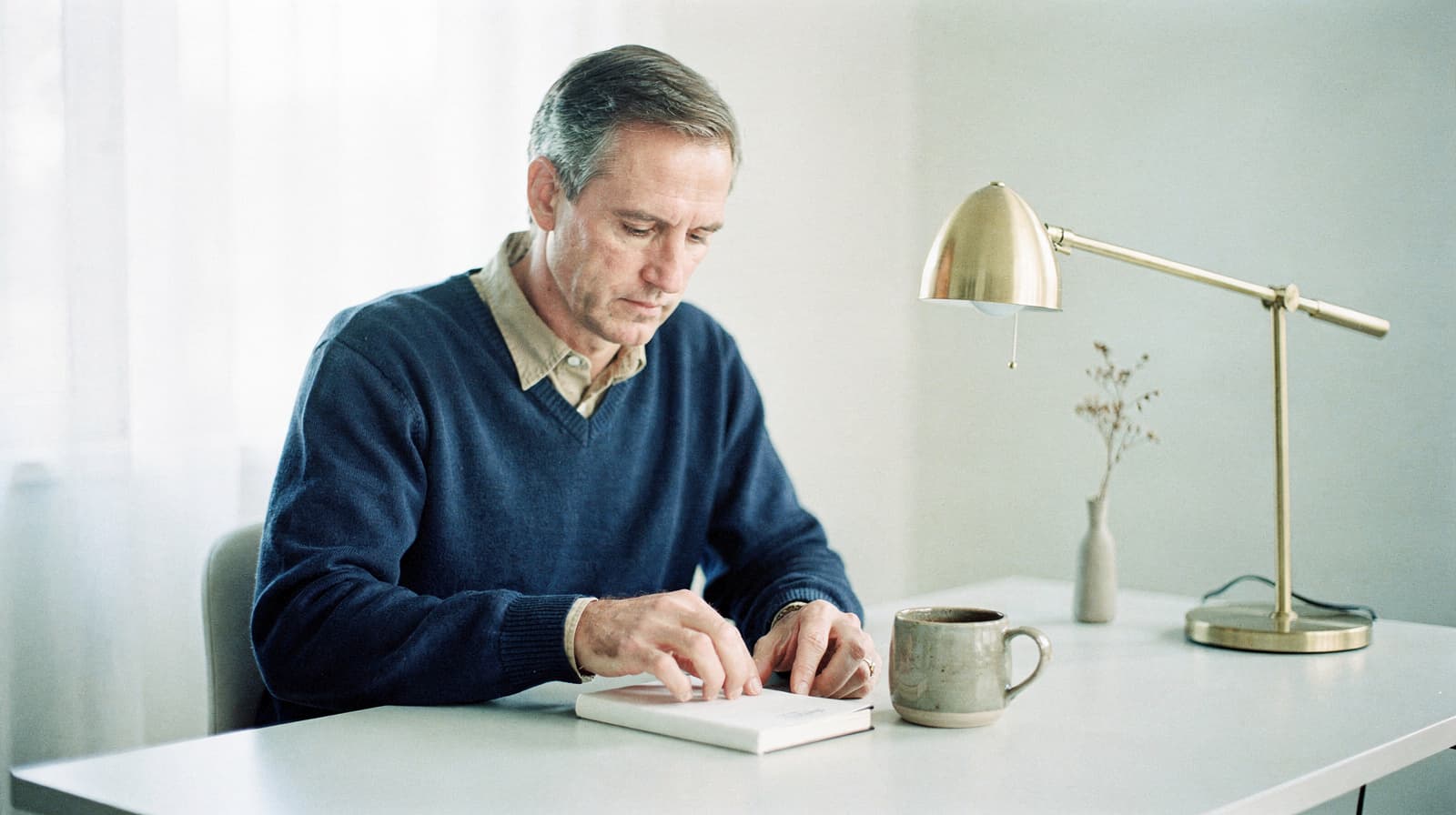 A man sits at a desk reading a book with a mug and a lamp nearby.