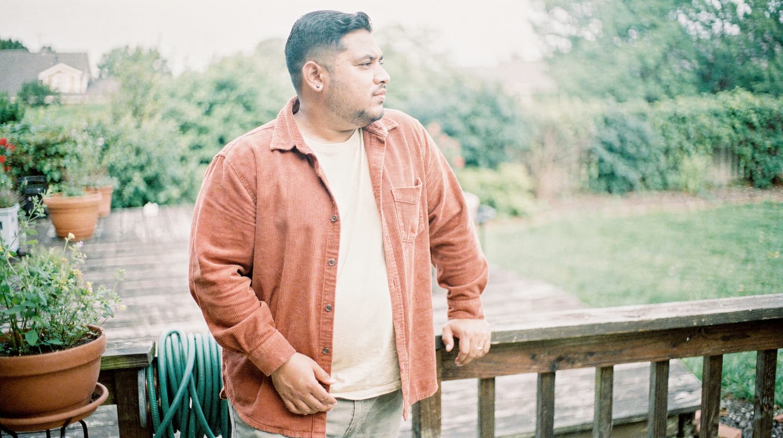 A man in a rust-colored jacket stands on a wooden deck, looking out over a garden with potted plants and greenery.